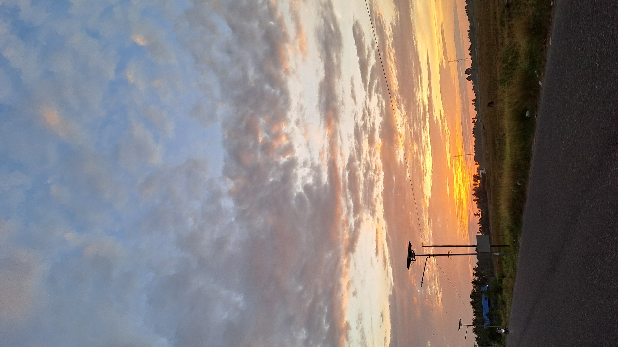 This image depicts a sunset in Cotgapu, Indonesia, over a rural landscape. The sky is filled with a dramatic display of clouds, tinged with hues of orange, pink, and blue from the setting sun. In the foreground, a paved road stretches into the distance, bordered by dry grass and vegetation.
Along the side of the road, two solar-powered streetlights stand tall, one of which is more prominent. In the middle ground, a line of trees, likely palm trees, forms a treeline with some buildings, including one with a blue roof, nestled amongst them. Further in the background, a vast field of harvested crops or dry grass extends to the horizon, dotted with a few silhouetted animals, possibly cows. Two tall, thin communication towers are visible in the distance on the right side of the frame. A single motorcycle with its headlight on is traveling on the road in the left portion of the image. The overall impression is one of tranquility and the natural beauty of the Indonesian countryside at dusk.