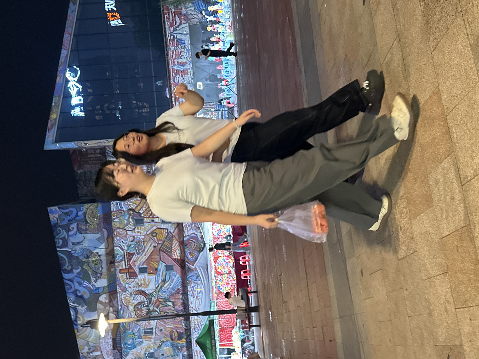 The image captures a nighttime street scene in the Chongqing High-tech Zone, China. Two young women are walking in the foreground, with the woman on the left carrying a clear plastic bag filled with orange-colored items. Both are wearing casual clothing and glasses. In the background, a building with bright, colorful murals and illuminated signage is visible. Further back, there appear to be market stalls with various goods and seating areas, suggesting a lively commercial or entertainment district. The ground is paved with stone tiles.