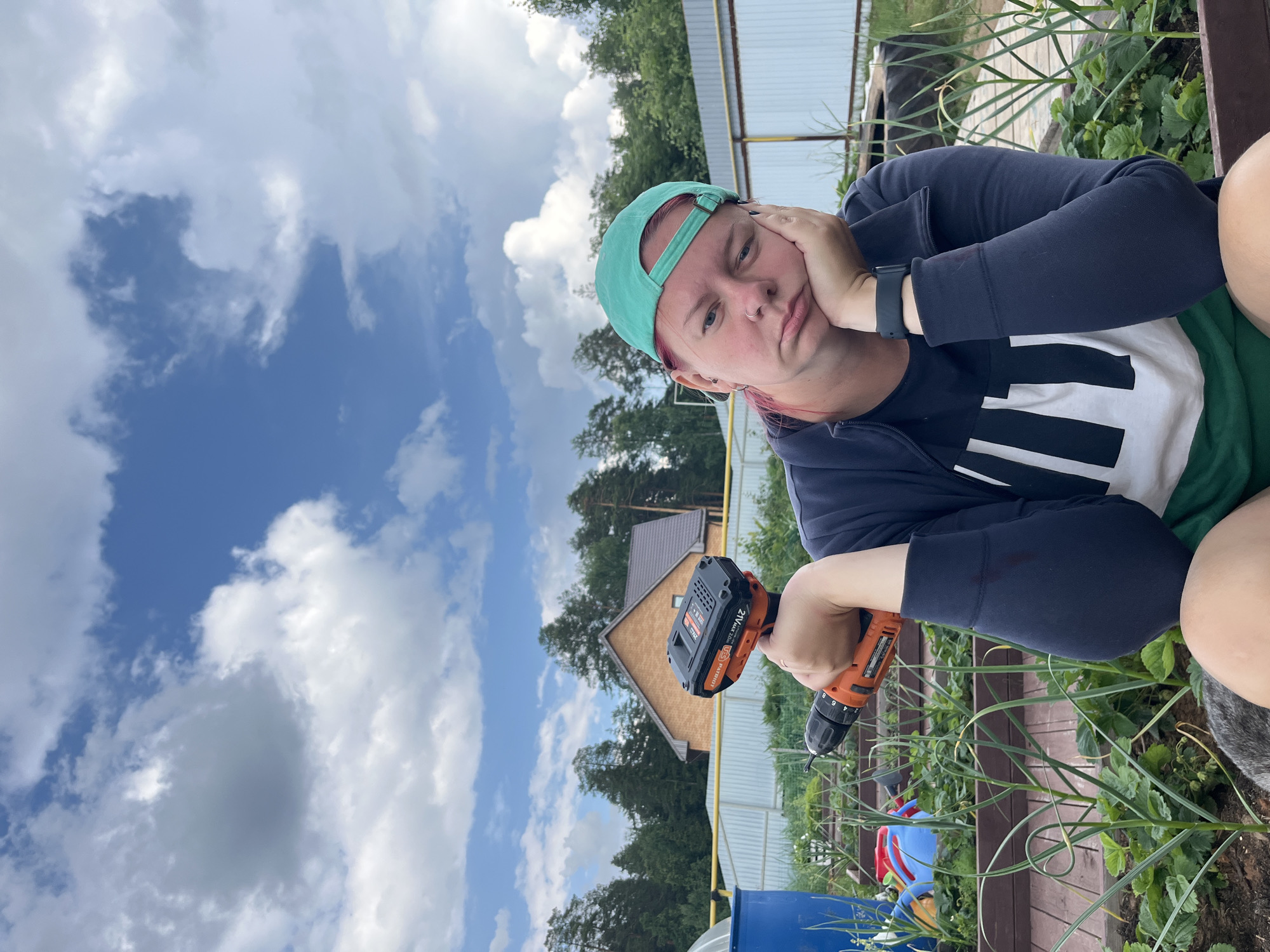 The image depicts a person sitting in a garden with a cordless drill in their right hand, held up and slightly out of frame. The person is wearing a green baseball cap, a dark blue zip-up hoodie over a white shirt with black stripes, and green shorts. Their left hand is propping up their face, and they have a bored or tired expression. In the background, there are raised garden beds with green plants, a fence, trees, and a brick house. The sky is blue with scattered white clouds. The overall scene suggests an outdoor setting, likely a backyard or a dacha (summer house garden) in a rural or suburban area. The city of Chaikovsky, Russia is located in a region with many such dacha settlements, which are popular for gardening and outdoor recreation.