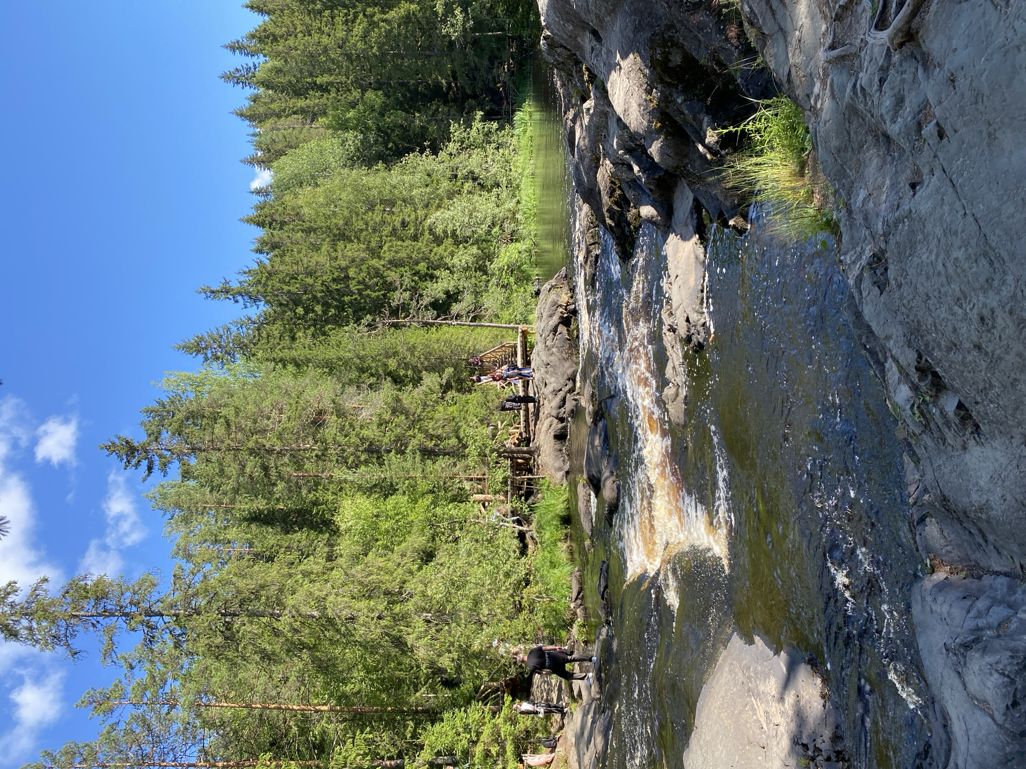 This image depicts a scenic view of the Ruskeala Mountain Park in Karelia, Russia, characterized by a natural landscape of coniferous forests, a river with rocky banks, and clear blue skies. In the foreground, the turbulent, shallow river flows over dark, uneven rocks, with patches of lighter, sandy-colored water indicating a small rapid. The riverbanks are lined with large, grey, textured boulders.
Midground features a wooden walkway or bridge structure spanning the river, with several individuals visible on or near it. A person in a black t-shirt and dark pants stands on the rocks on the left bank, facing away from the camera and towards the river. Further along the left bank, a few other individuals are seen walking. On the right bank, more people are observed on the rocks and near the wooden structure.
The background is dominated by a dense, green coniferous forest that extends to the horizon under a bright blue sky with a few wispy white clouds. The overall impression is of a natural recreation area during daytime, with visitors enjoying the outdoor environment.