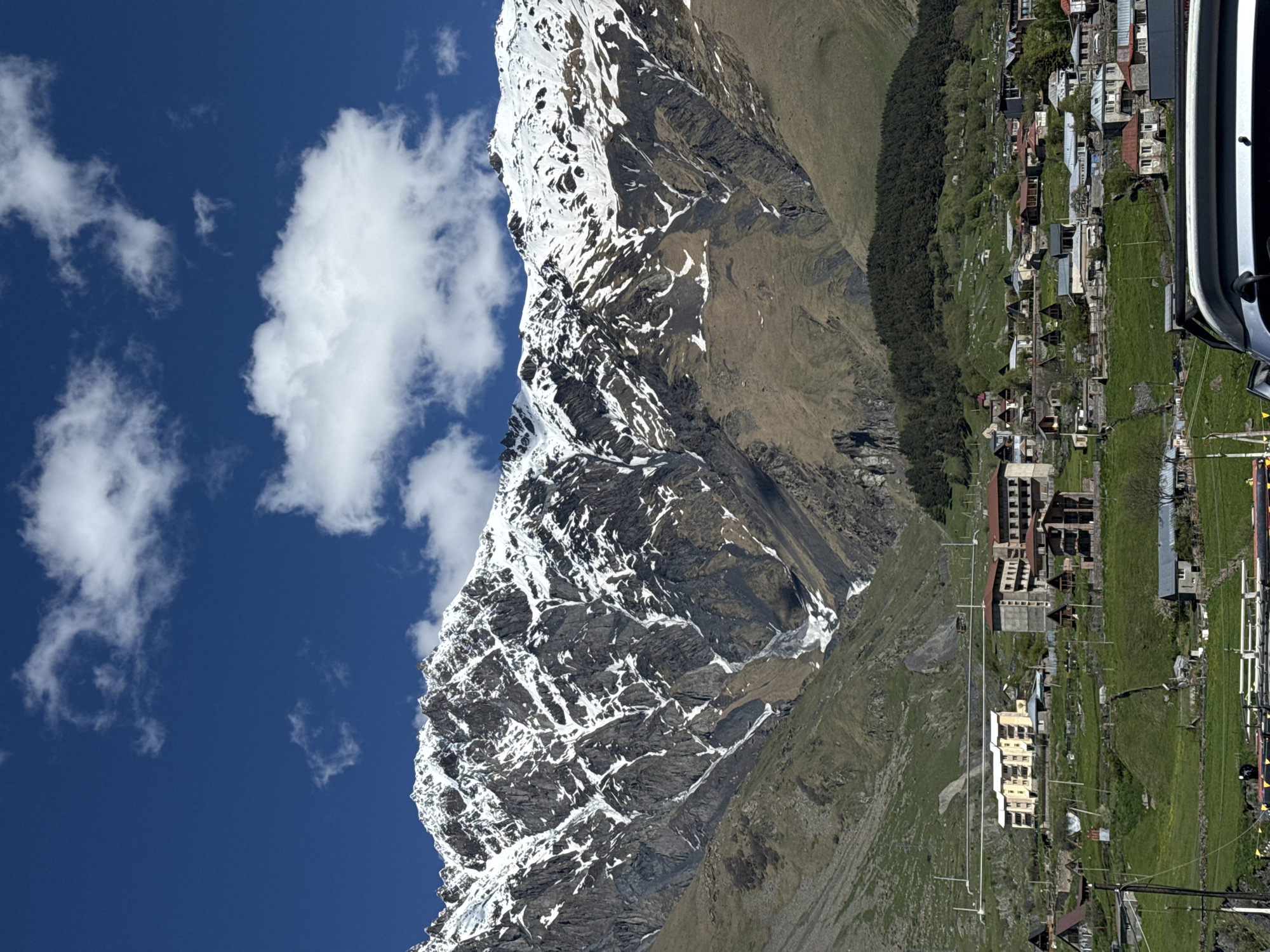 The image presents a vertical view of a mountainous landscape with a clear blue sky and scattered white clouds. A snow-capped mountain dominates the left side of the image. The mountain features rocky terrain with patches of snow and ice.
To the right of the mountain is a green, grassy hillside that descends into a valley. The village of Stepantsminda is nestled in the valley, consisting of buildings with light-colored exteriors and red or gray roofs. Some buildings are larger and appear to be hotels or apartment complexes. A dark green forest line runs along the upper edge of the valley.
In the foreground, on the right side of the image, part of a vehicle is visible. A grassy field stretches out in front of the vehicle, and power lines run through the village. The photograph is taken from an elevated position, looking down on the village and surrounding area.