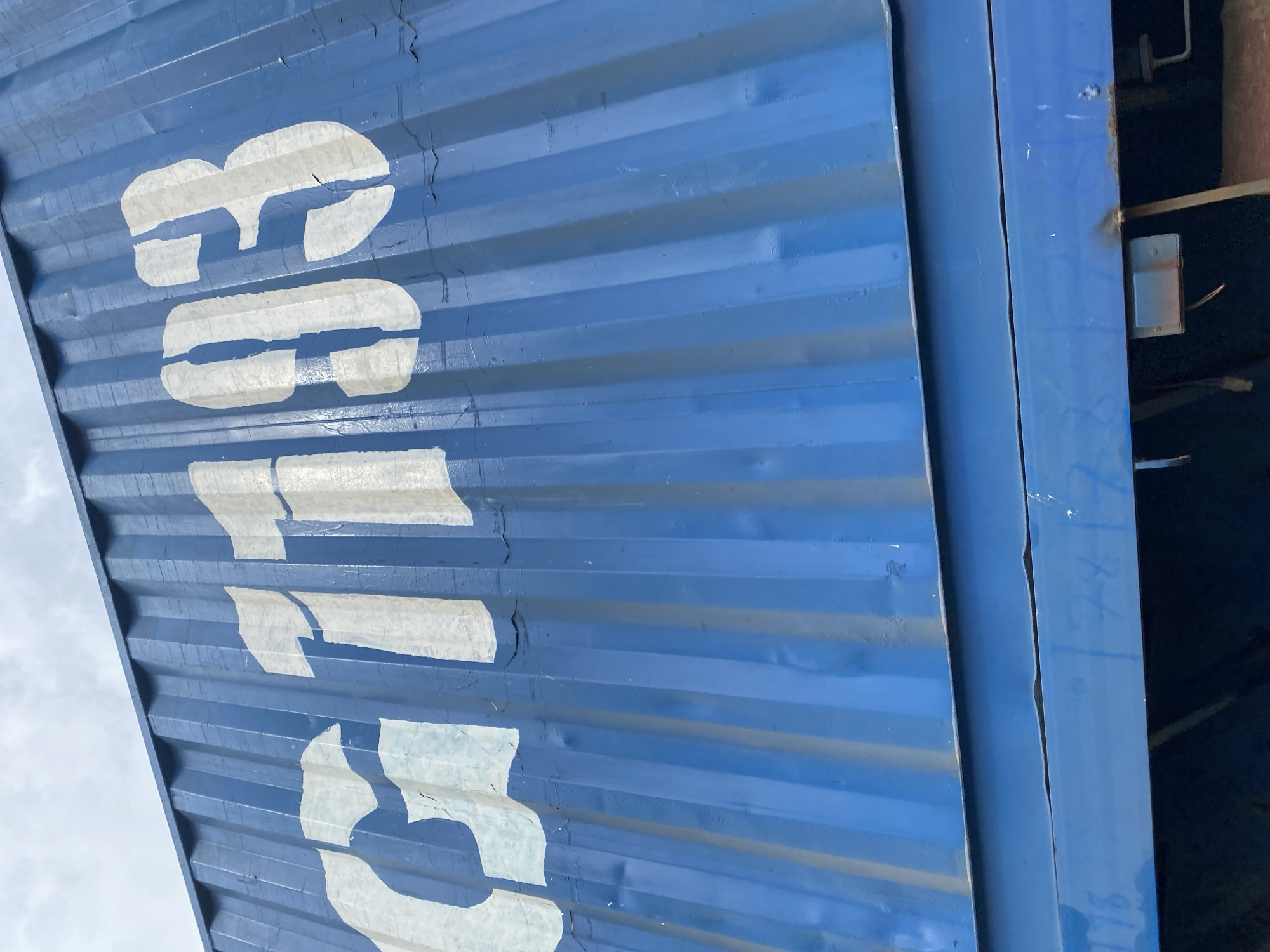 The image shows the side of a blue shipping container with the number "G103" stenciled in white. The container is ribbed and appears weathered, with some cracks and peeling paint visible. The sky above is overcast. The bottom of the image shows part of another blue structure, possibly a trailer or another container, with some faint markings or numbers that are difficult to discern. A small rectangular metal box with wires is attached to this lower structure. The scene appears to be in an outdoor industrial or transportation yard, possibly in the city of Kuru, Nigeria, given the context provided. There are no people or animals visible in the image.
