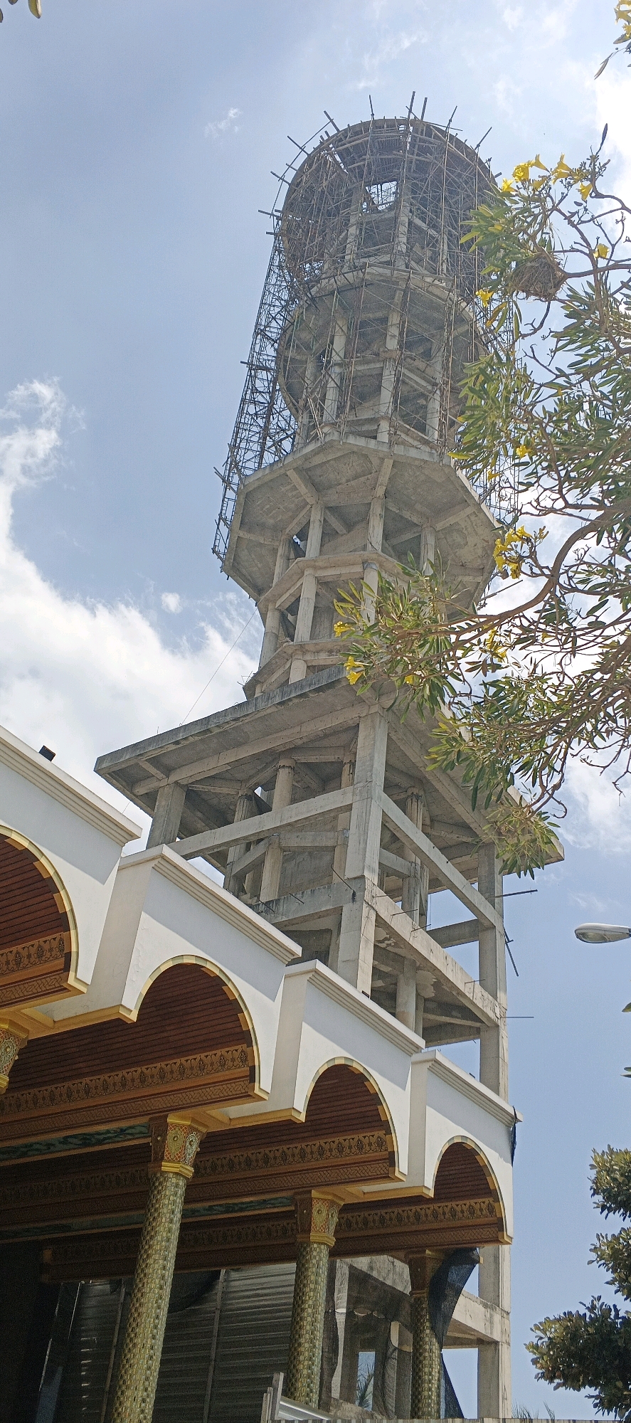The image shows a partially constructed tower in the city of Kandangsemangkon, Indonesia. The tower is made of concrete and scaffolding, with a circular top section. In the foreground, there is a building with arched windows and decorative pillars. A tree with yellow flowers is visible on the right side of the image. The sky is blue with some clouds. There are no people visible in the image.