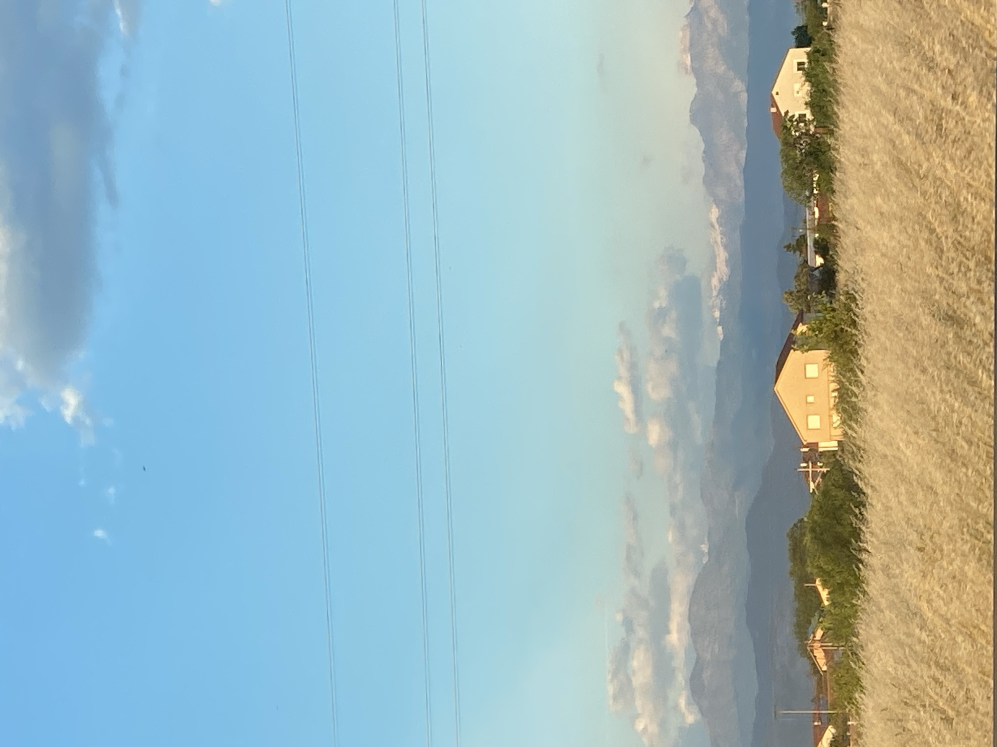 The image shows a landscape in Mahala, Montenegro. The scene includes a light blue sky with clouds on the left. Power lines stretch vertically across the left side of the frame. In the center, there are distant mountains with interspersed clouds. On the right, there are houses with tan exteriors, separated by trees. The foreground on the far right features tall, dry grasses.