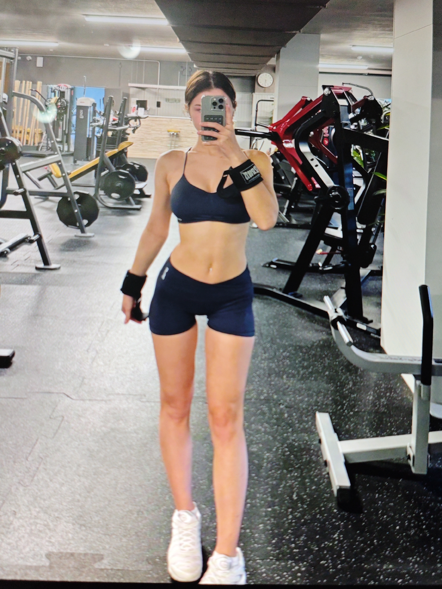 A full-body, front-facing selfie is taken in a gym located in Baku, Azerbaijan. The subject, a woman with brown hair pulled back, is wearing a dark blue sports bra and matching shorts. She has on black wrist weights. Her hair is pulled back into a bun. She is holding a smartphone in her right hand, which is angled to capture her reflection. The gym environment features various weightlifting machines, including a leg press and a smith machine, along with weight plates and benches. The floor is covered with black rubber mats. In the background, a large round clock is visible on the wall.