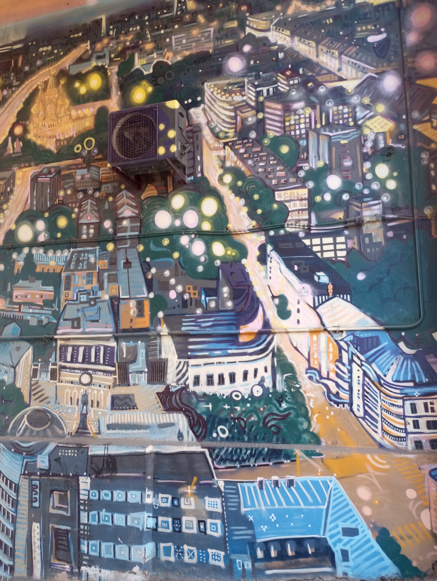 The image shows a painted mural on a building exterior in Pervomaysky, Russia. The mural depicts a stylized, densely packed cityscape at night, with various architectural styles mixed together. The city is rendered with blues, greys, yellows, and greens. There are circular shapes scattered throughout that may represent light sources like streetlights or stars. The foreground appears to show buildings at a lower level while the background features structures higher up, creating a sense of depth. An air conditioning unit is attached to the wall, partially obscuring a section of the mural. The mural is painted on a surface that shows signs of wear, with cracks and uneven textures visible.