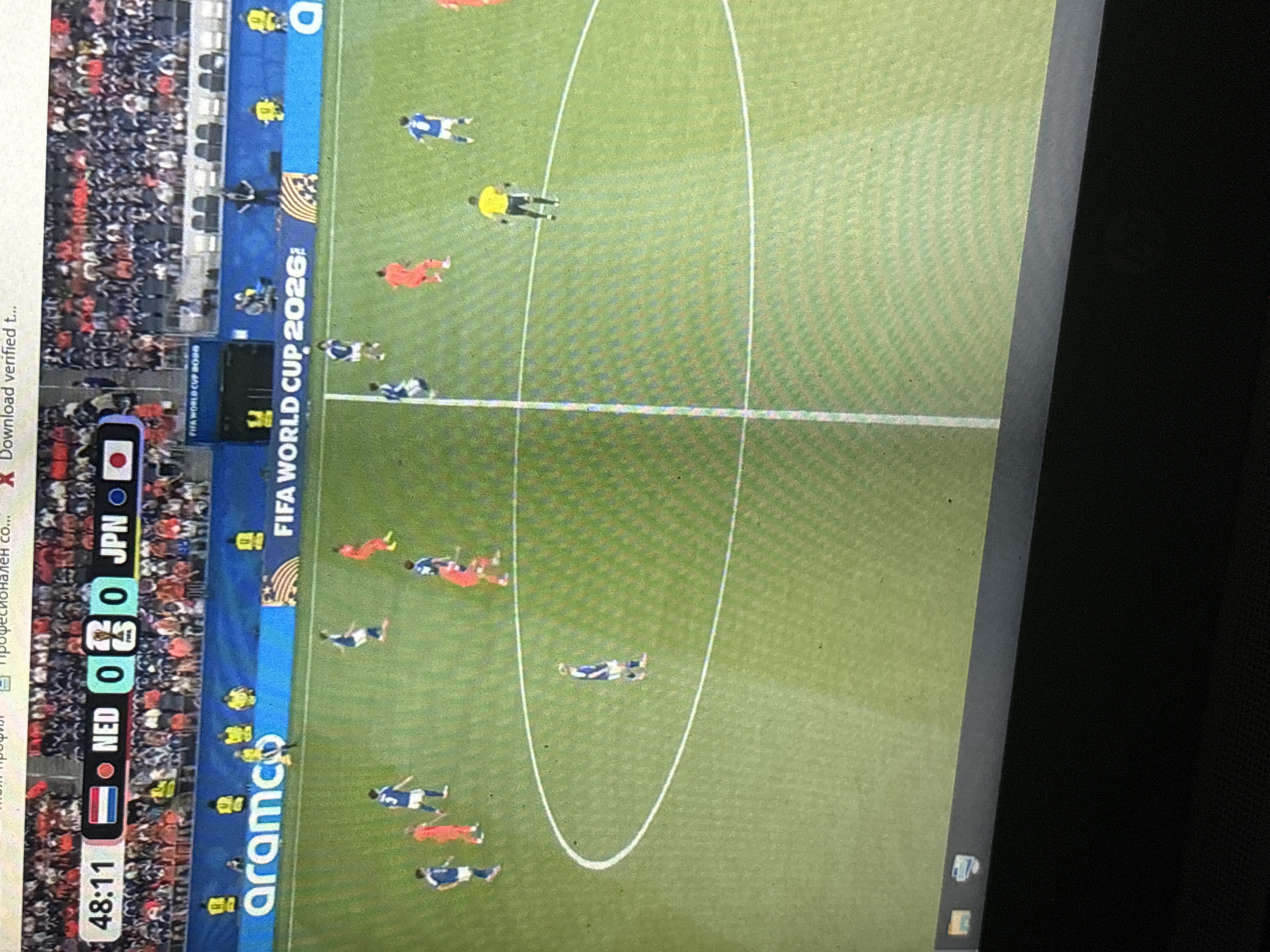 The image is a photograph of a soccer match displayed on a screen, possibly a laptop or television. The screen shows a live broadcast with a score overlay indicating the time is 48:11, with the score being NED 0 - 0 JPN. Flags representing the Netherlands (NED) and Japan (JPN) are visible. A logo for "arama" is displayed on a blue banner in the background, along with "FIFA WORLD CUP 2026". The soccer field is green with white lines. Several players in blue and orange jerseys are visible on the field, along with a referee in a yellow jersey. The image appears to be taken from an angle, with the bottom portion of a dark surface, likely the device the image is displayed on, visible in the foreground. The scene is indoors or in a controlled environment where the screen is being viewed. There is no discernible connection to the city of Enevo, Bulgaria.