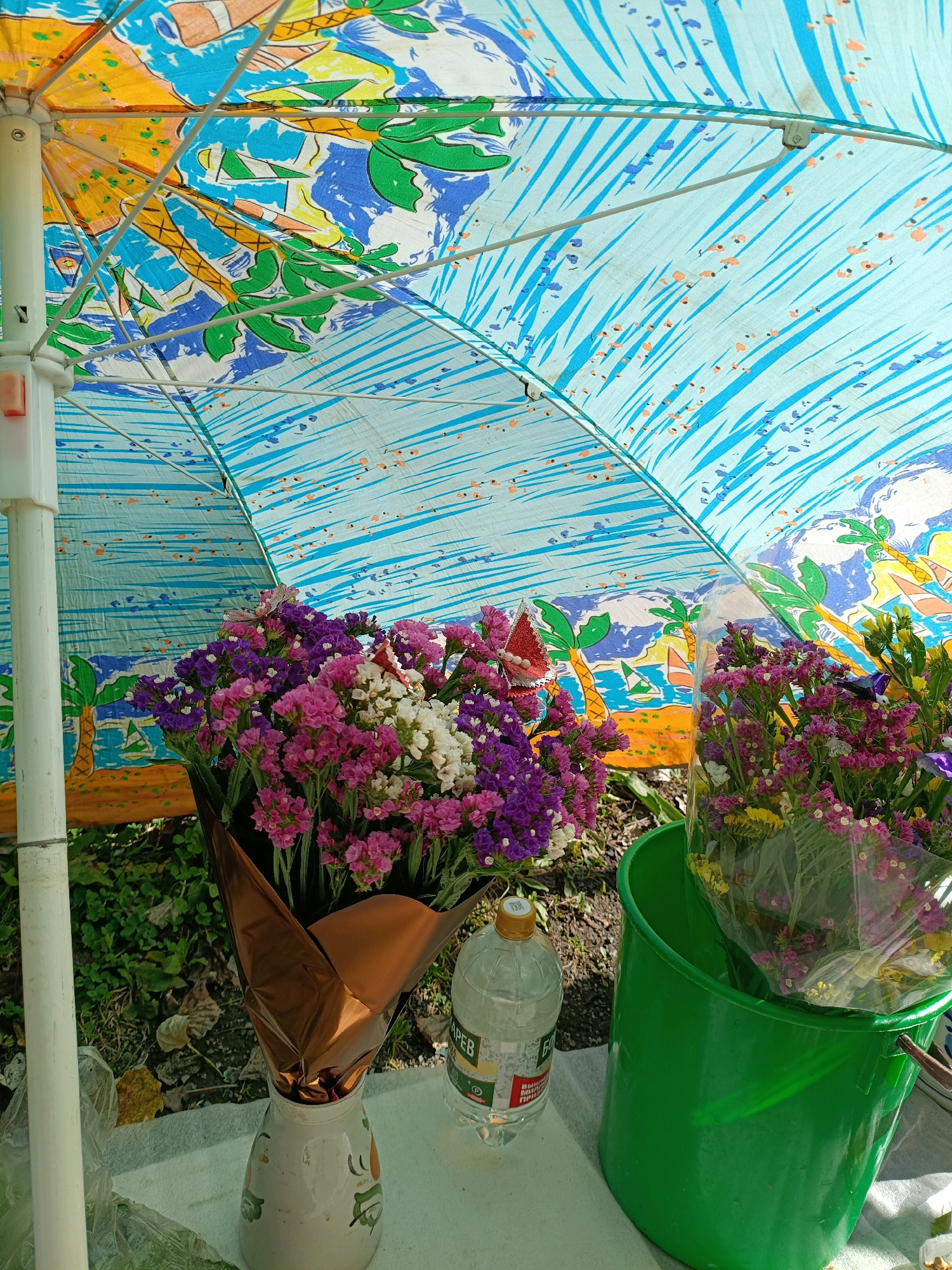 The image depicts a close-up view of a colorful, open umbrella with a tropical beach scene printed on it, featuring palm trees, sand, and water. Beneath the umbrella, there are two bouquets of small, multi-colored flowers (pink, purple, and white). One bouquet is in a brown paper wrapper, placed in a white ceramic vase with green painted designs. The second bouquet is wrapped in clear plastic and sits inside a bright green bucket. A clear plastic water bottle with a yellow cap is positioned on the ground next to the bouquets. The ground is covered with a white cloth or tarp, with visible grass and fallen leaves in the background. The scene suggests an outdoor market or street vendor stall, likely in the city of Asbest, Russia, given the presence of a water bottle with Russian labeling. There are no people visible in the image.