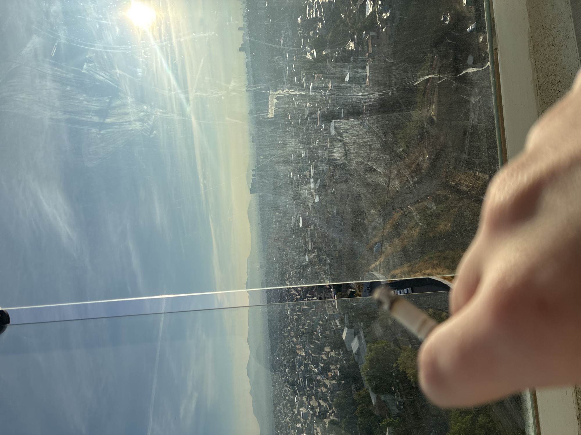 The image shows a view of a cityscape, believed to be in Huixquilucan de Degollado, Mexico, seen through a dirty window. In the right foreground, a blurred human hand holds a lit cigarette or joint. The city spans a vast area with visible buildings and vegetation. The sky is visible through a portion of the window. The window itself appears scratched and smudged, impacting the clarity of the cityscape.