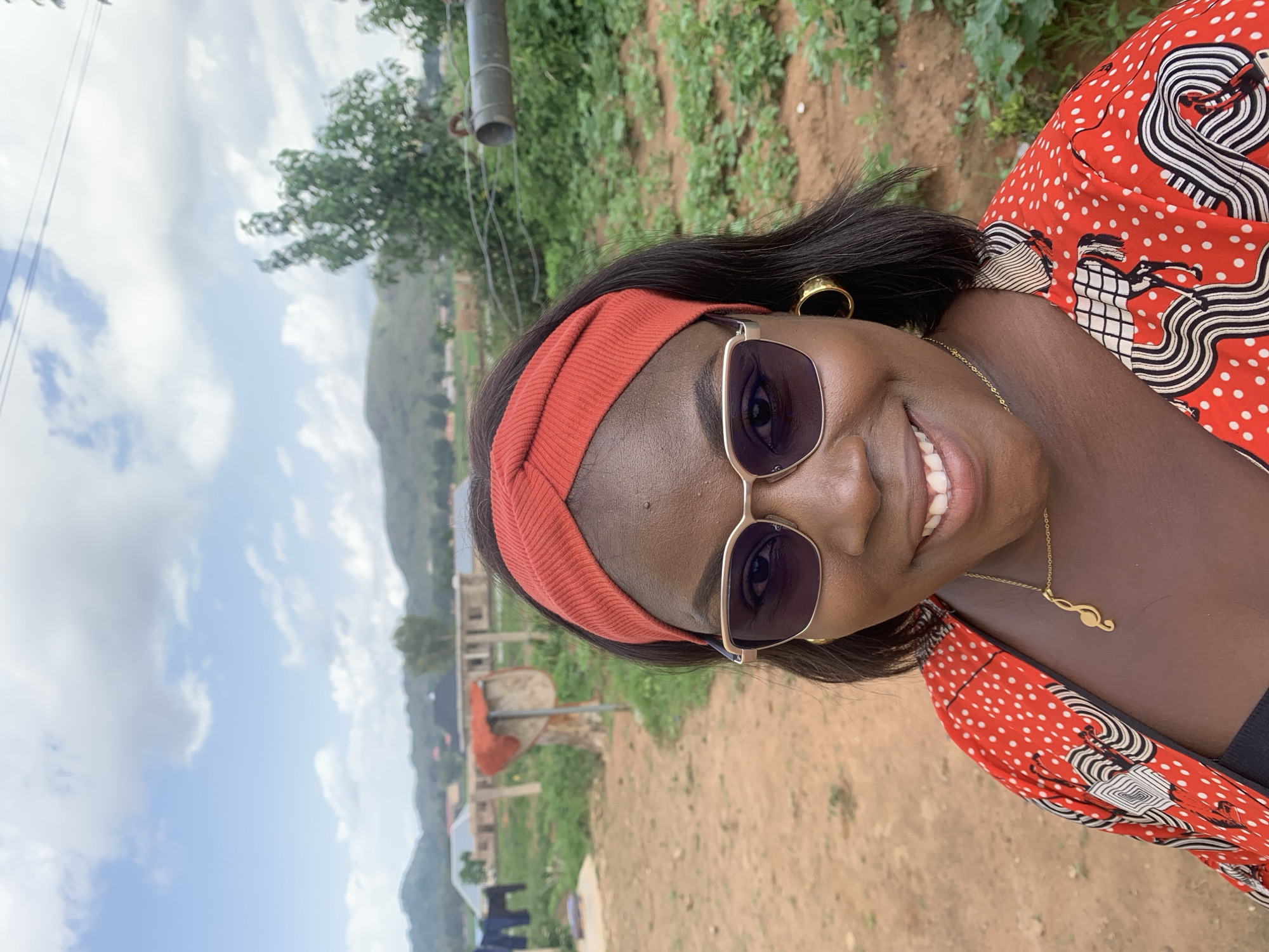 A medium shot shows a Black woman in Kuru Karama, Nigeria, smiling at the camera. She is wearing a red and white patterned top, a red headband, and gold hoop earrings. She also has on sunglasses and a gold necklace with a music note pendant. The background shows a rural landscape with rolling hills, trees, and a few buildings. There is also a clothesline with laundry hanging on it. The sky is blue with scattered clouds.