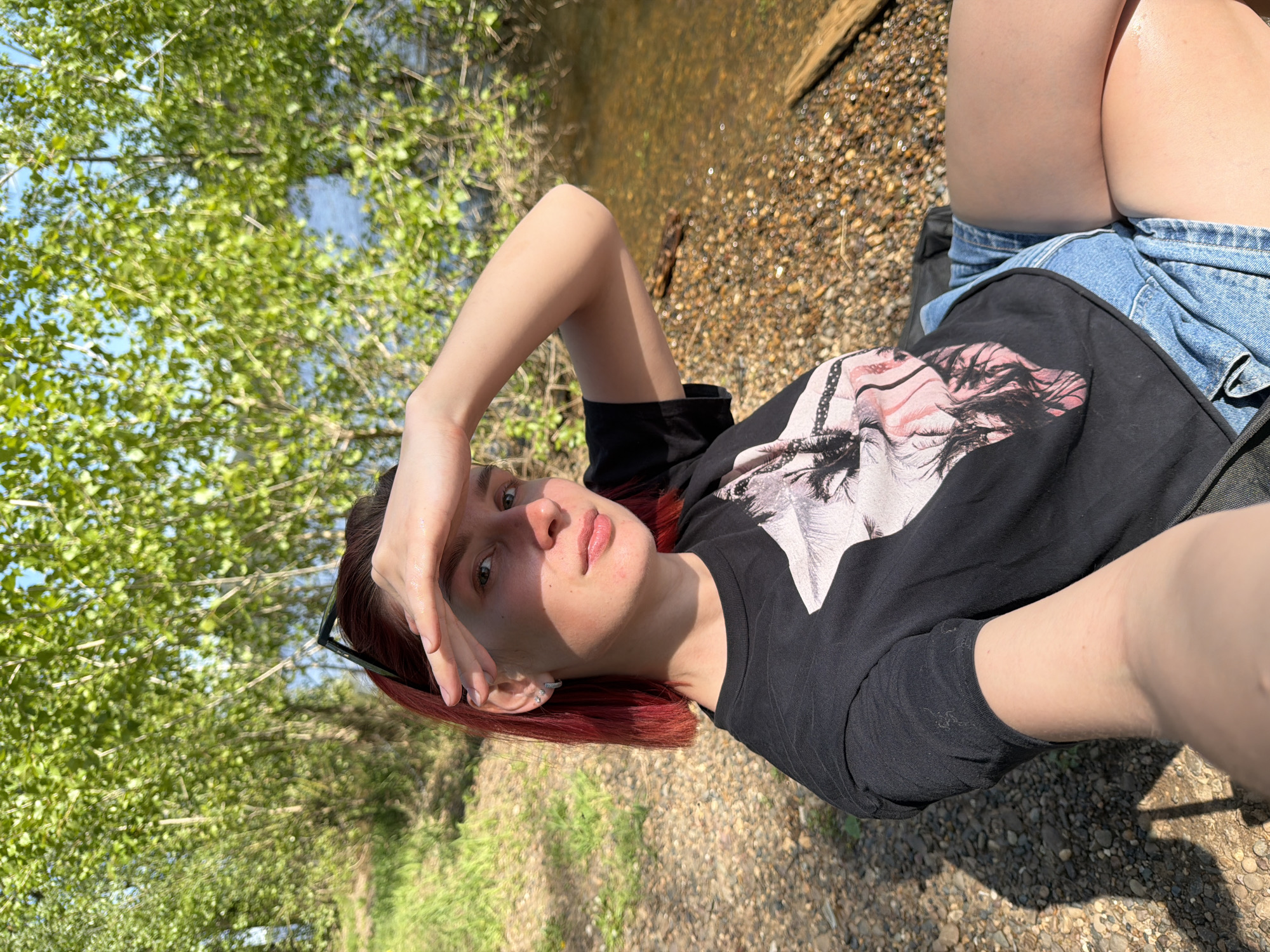A medium shot shows a fair-skinned person lying outdoors in a field of pebbles or small rocks, with trees and water visible in the background. The person, likely female, has short red hair and wears a black graphic T-shirt, along with blue jean shorts. Her left arm is raised, with her hand shielding her eyes. Sunglasses are tucked behind her ear. The photo appears to be taken in daylight with a wide-angle lens. The location is not identifiable as Noviy, Russia, from the details in the image.