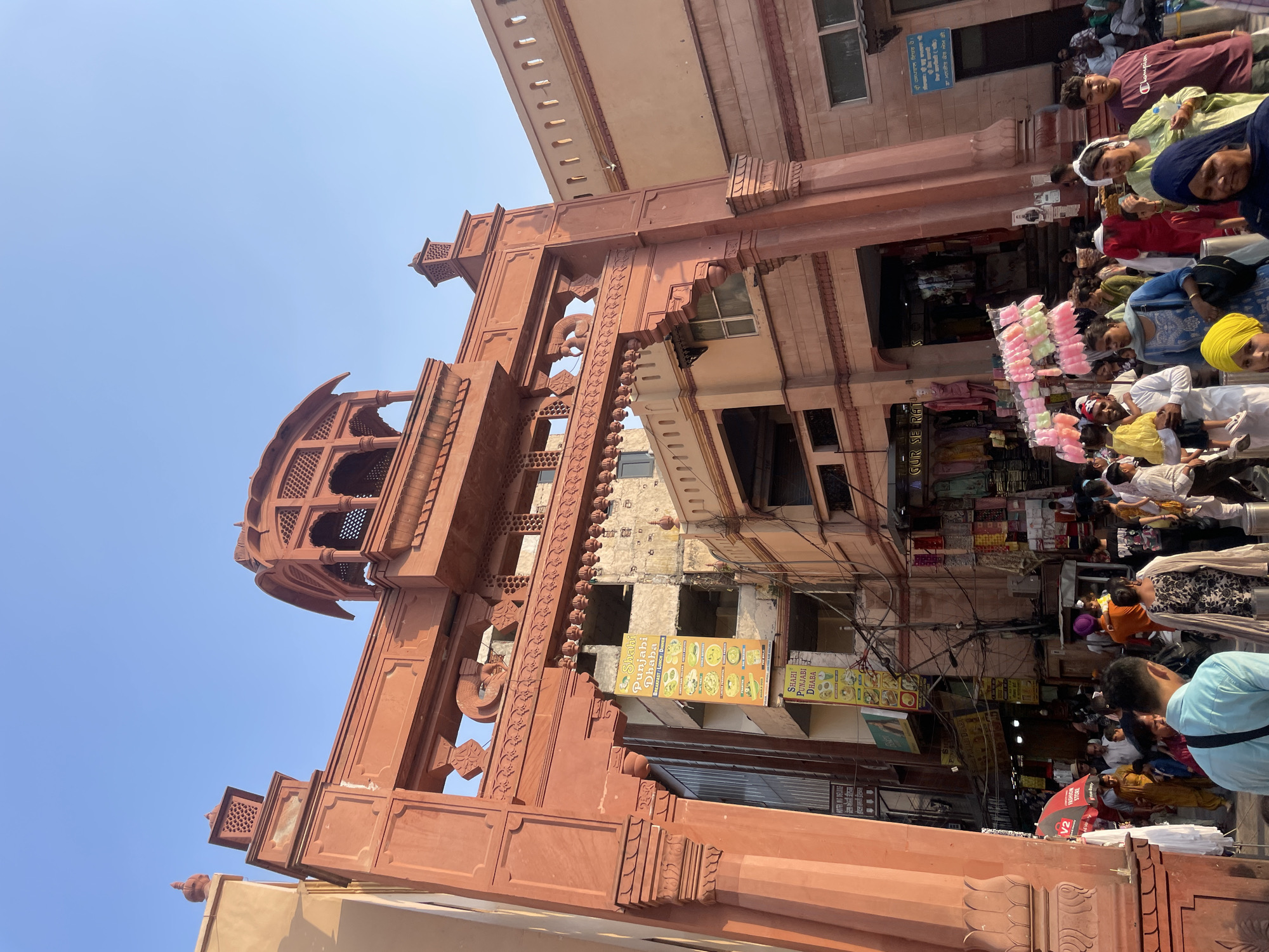 The image depicts a bustling street scene in Amritsar, India, likely near the Golden Temple. In the foreground, a large, ornate red sandstone archway dominates the frame, adorned with intricate carvings and decorative elements. The archway frames a view of a narrow street lined with shops and buildings. A crowd of people are walking through the archway and along the street, some dressed in traditional Indian attire. Several shops are visible, displaying a variety of goods, including clothing, food, and other merchandise. Signs in Punjabi are visible, advertising local businesses. The atmosphere appears lively and vibrant, with a sense of everyday life unfolding in this Indian city. The sky above is clear and blue.