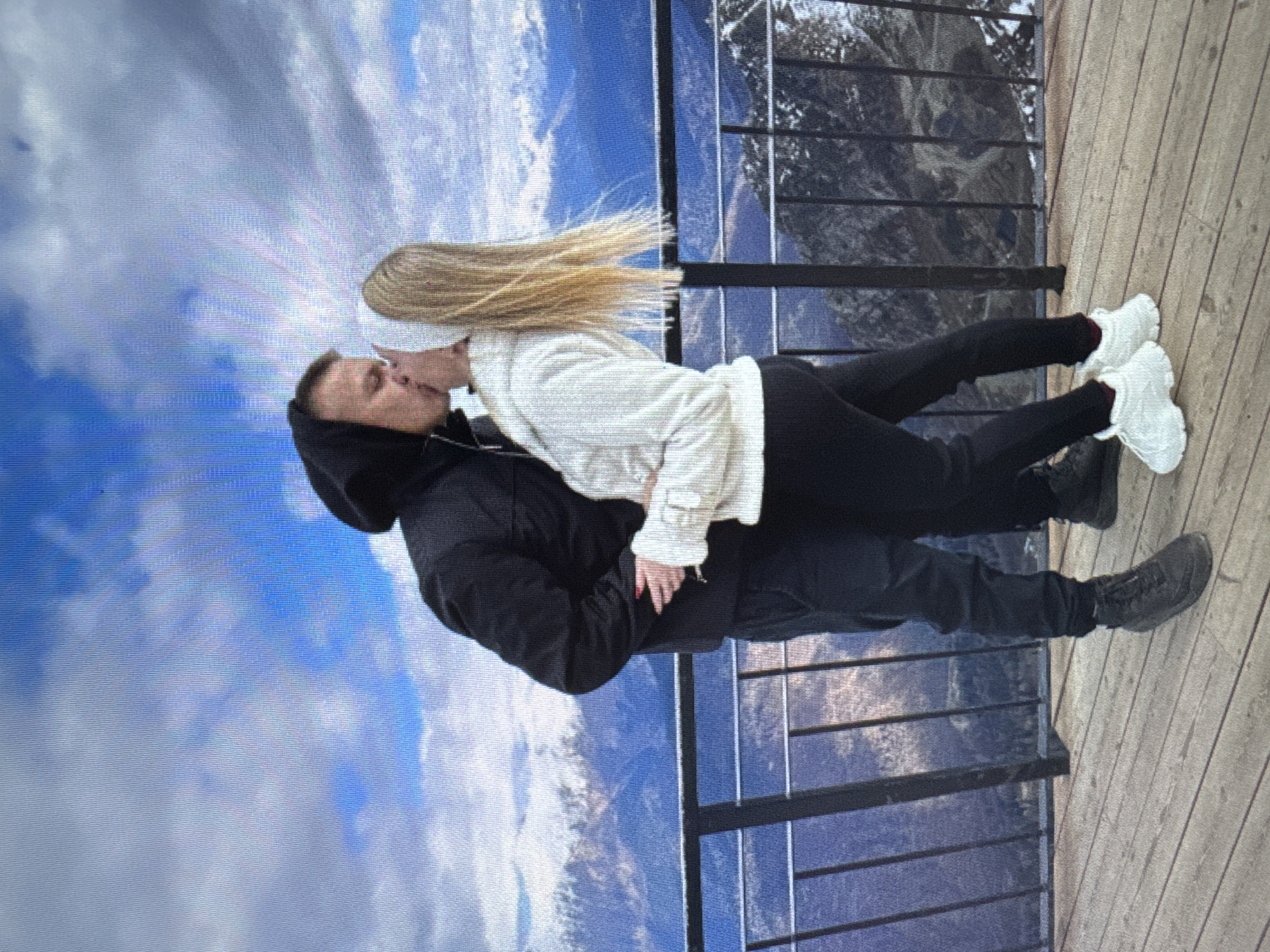 A couple is captured in a romantic moment, kissing while standing on a wooden platform overlooking a vast mountain range under a partly cloudy sky. The man, on the left, wears a black hooded jacket and black pants, with dark sneakers. The woman, on the right, sports a white hoodie and black leggings, paired with white chunky sneakers. They are positioned at a railing, with the dramatic mountainous landscape serving as their backdrop. The image suggests a scenic viewpoint, possibly in a mountainous region.
