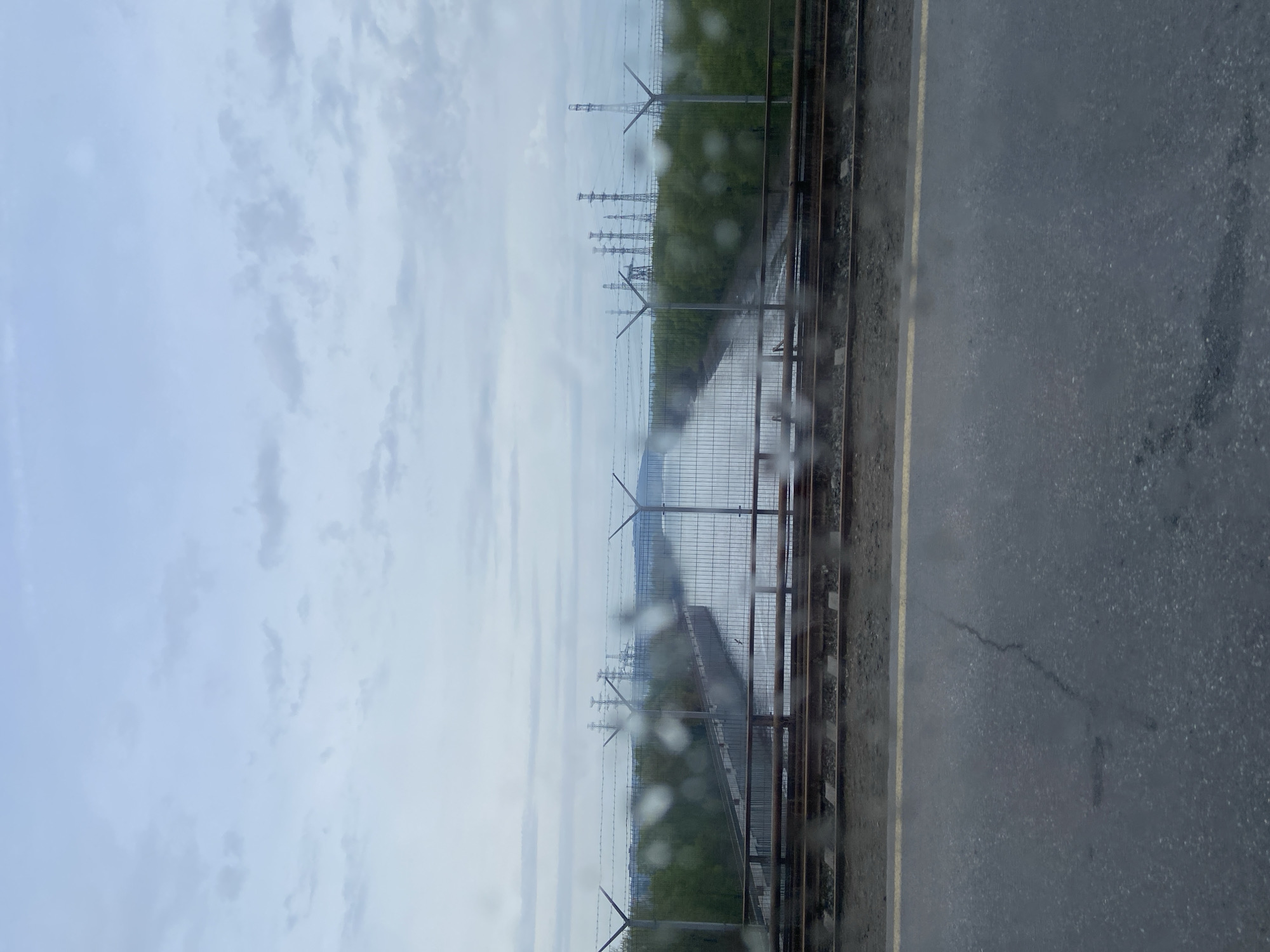 The image, taken through a rain-streaked window, shows a view towards the city of Chaikovsky, Russia. Visible is a gray sky with scattered clouds. The foreground features railway tracks and a gray concrete surface with a yellow line. A fence topped with barbed wire runs parallel to the tracks, and in the distance, power transmission towers are visible, partially obscured by trees. The environment appears industrial or peri-urban. No people are apparent in the scene.