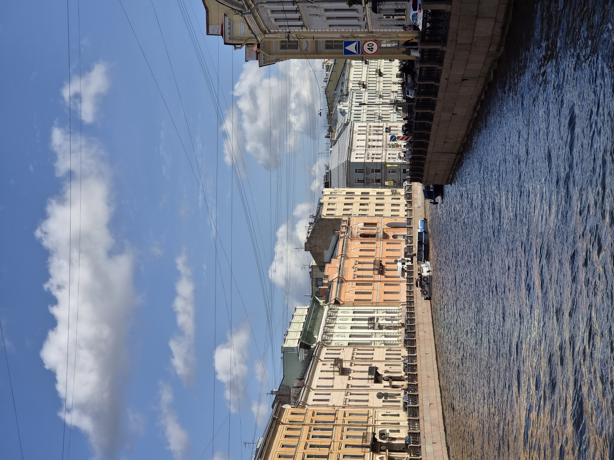 This is a wide shot of a canal bordered by ornate buildings. The buildings are painted in various pastel colors, including yellow, peach, and cream, and feature classical architectural details. Many have balconies and arched windows. The canal water is dark blue and slightly choppy. Several boats are visible on the water, including a white tour boat and a dark blue vessel. A stone embankment with a decorative metal railing lines the canal. On the embankment, people are walking, and cars are parked. Overhead, power lines crisscross the sky, which is a bright blue with scattered white clouds. A traffic sign with a speed limit of 40 is visible on the right side of the image. The overall scene depicts an urban European canal setting.