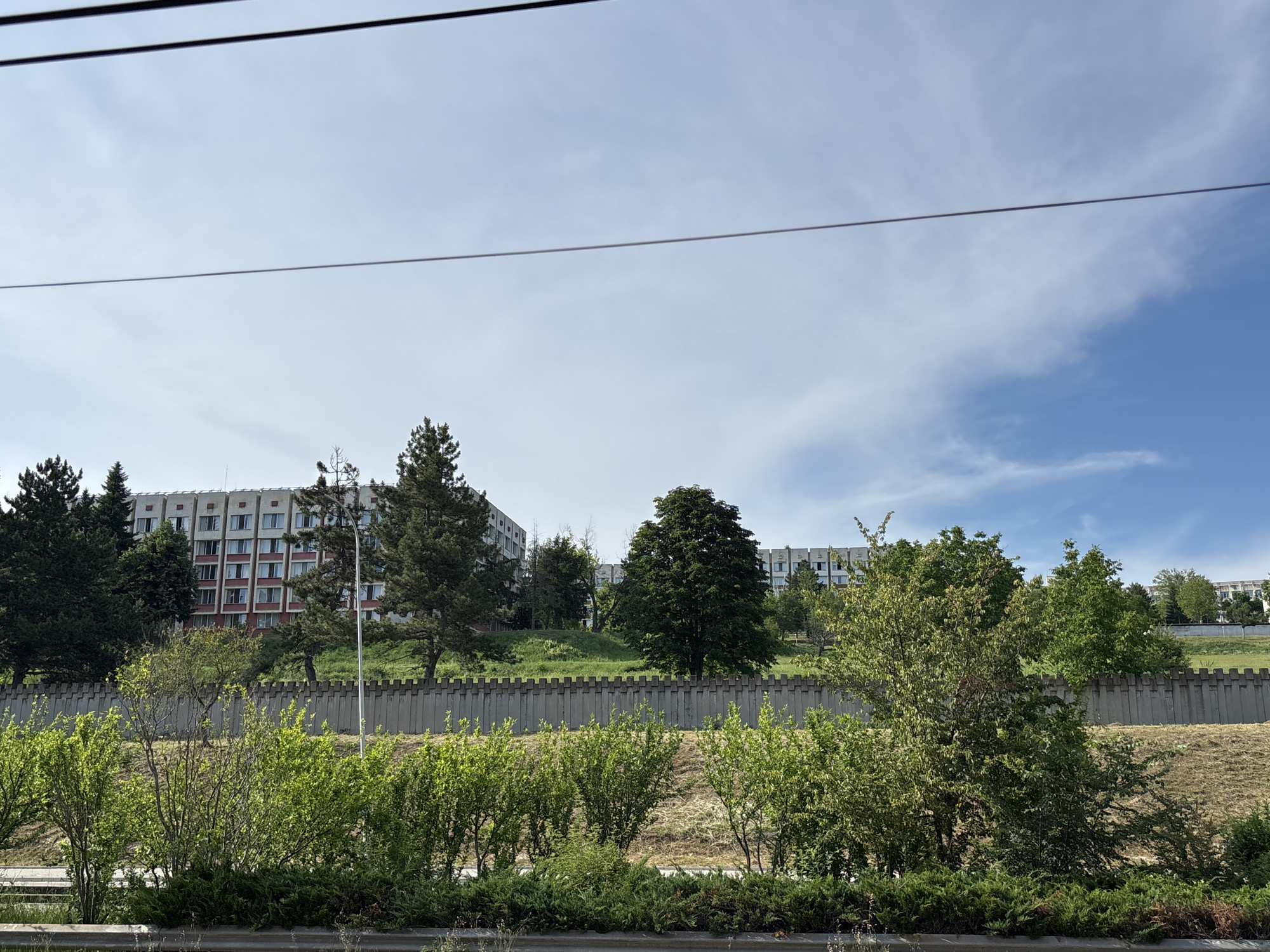 The image depicts a daytime scene in Veliko Tarnovo, Bulgaria, featuring a row of apartment buildings on a hill overlooking a concrete barrier and lush green vegetation in the foreground. The apartment buildings are multi-story structures with a light-colored facade and red accents around the windows. They appear to be of Soviet-era architecture. Several trees, including evergreens and deciduous trees, are scattered across the hillside between the buildings and the barrier. The concrete barrier is a textured, repeating pattern of rectangular blocks. In the foreground, dense bushes and smaller trees are visible, with power lines stretching across the top of the frame against a partly cloudy sky. The overall impression is of a residential area with a mix of urban architecture and natural elements.