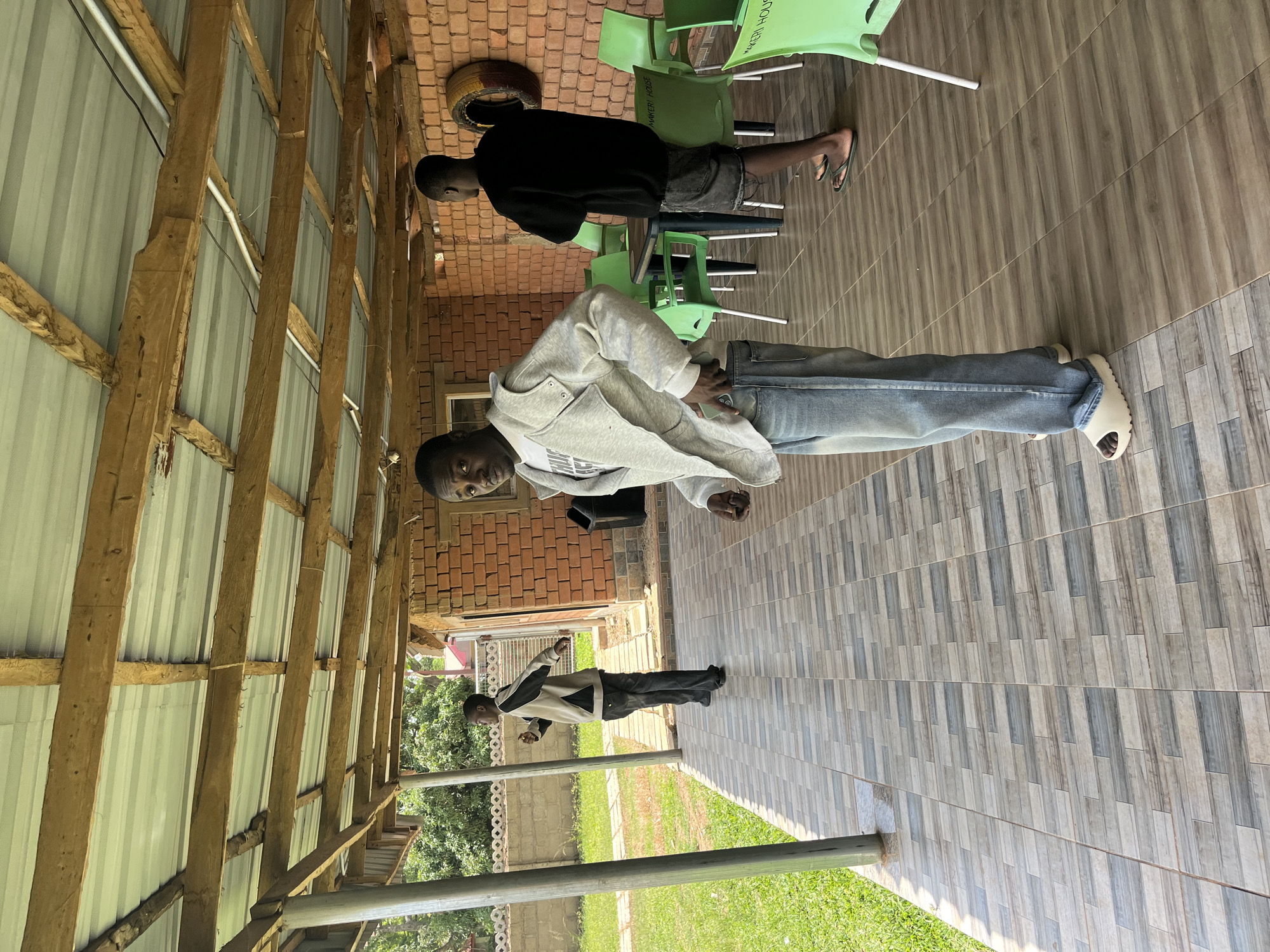 The image depicts an outdoor scene with three young Black men. One man, wearing a black t-shirt and shorts, is standing, possibly in a casual gathering or waiting. Another man, in a light grey hooded jacket and blue jeans, is standing and looking towards the camera. The third man, wearing a black and grey jacket and black pants, is standing and looking away from the camera. The setting appears to be a covered outdoor area with a wooden beam structure and a corrugated metal roof. The floor is tiled with a grey and brown pattern. In the background, there are brick walls and green chairs, suggesting a communal or outdoor seating area. A grassy area is visible beyond the tiled floor, with trees and foliage suggesting a temperate environment. The lighting suggests it is daytime. There is no visible text in the image.