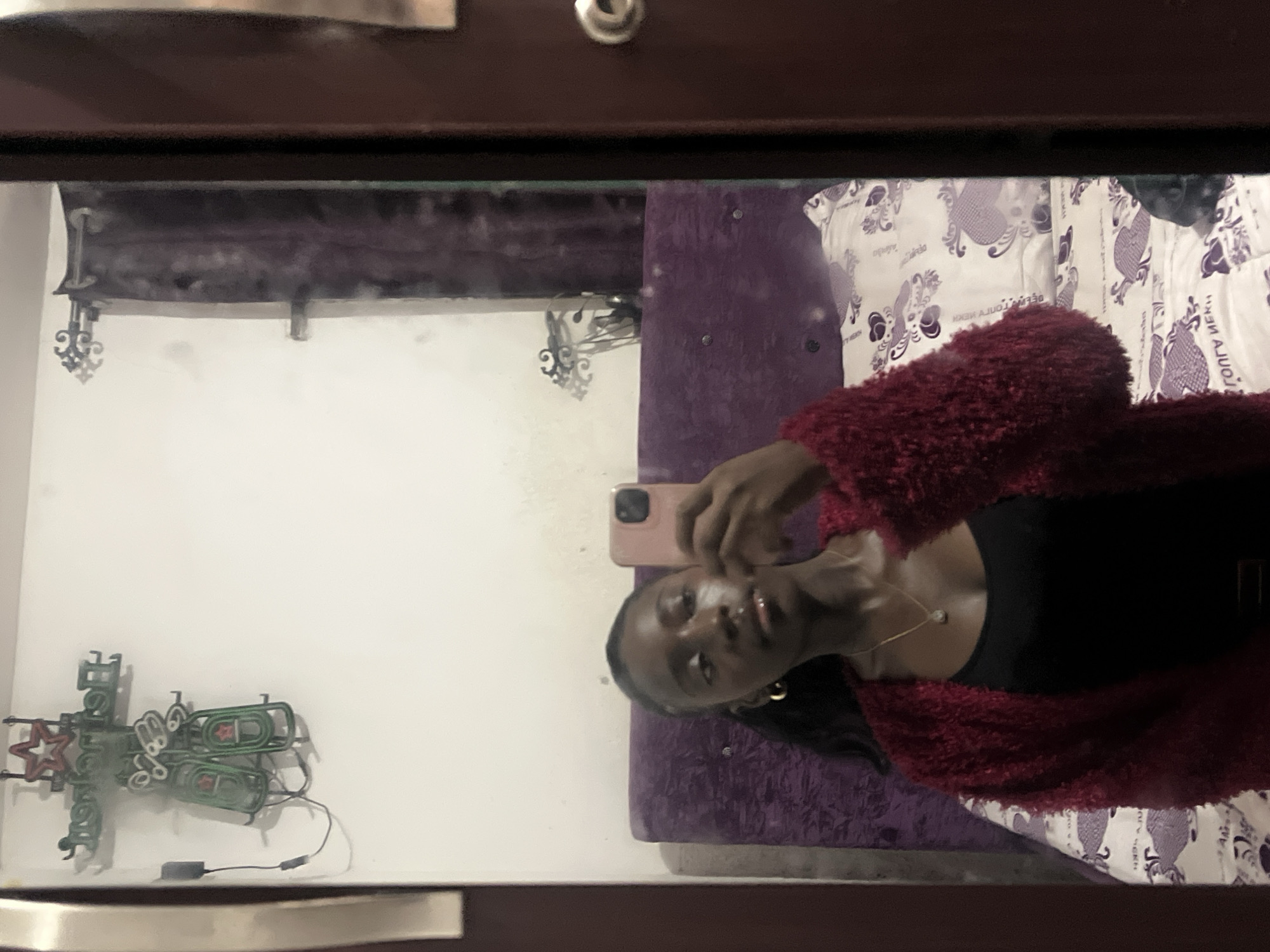 A woman is taking a selfie in a mirror inside a room. The mirror is framed by a dark brown wooden border. In the reflection, the woman is wearing a black top and a red fuzzy jacket. She holds a pink phone in her hand. Behind her is a purple bedspread and patterned white and purple pillows. On the wall opposite her in the reflection, there is a Heineken neon sign. The specific location within Abuja, Nigeria, cannot be determined from the image.