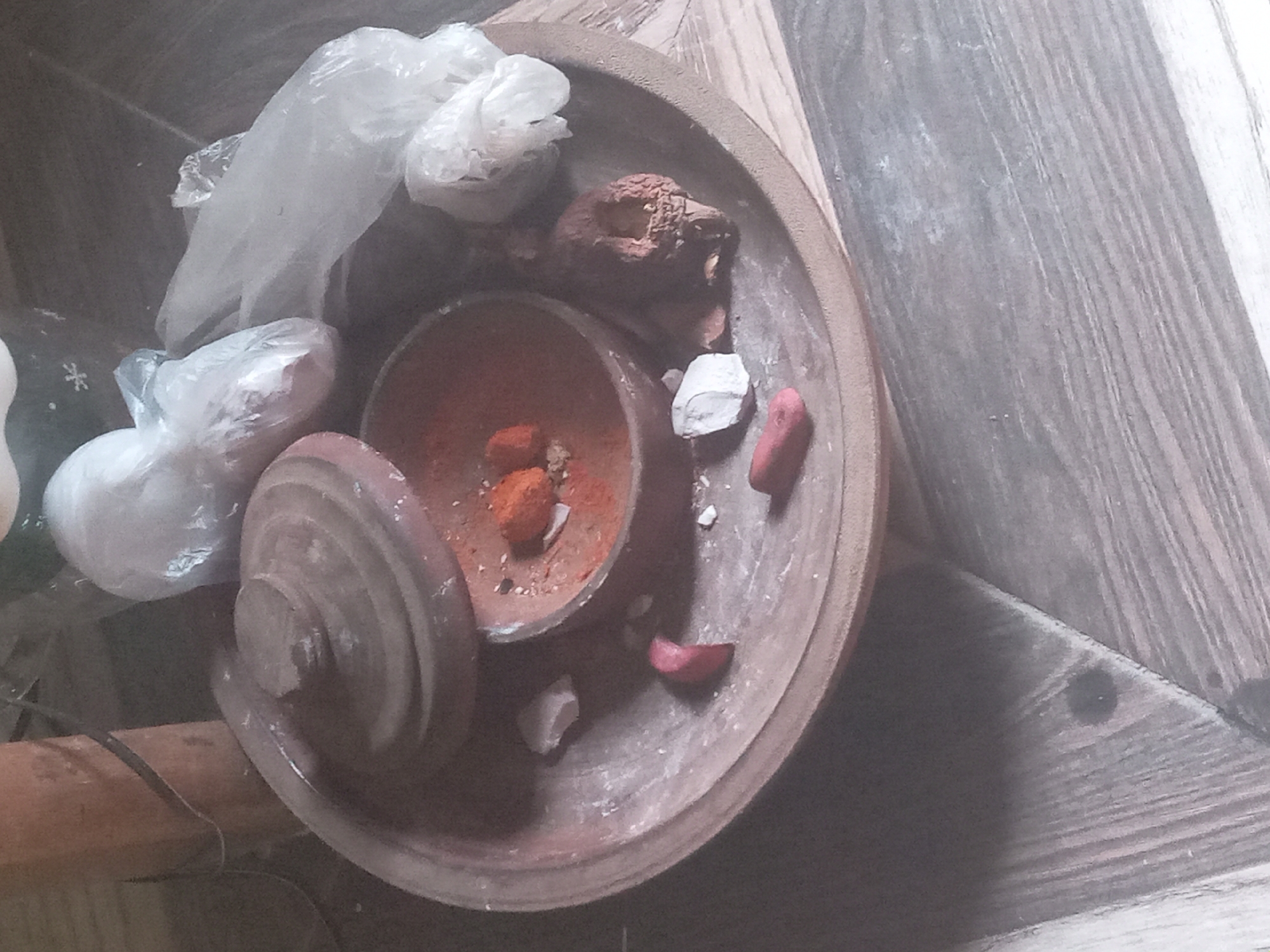 The image displays an overhead view of a collection of traditional materials arranged within a large, circular, terracotta-like tray, set upon a striped wooden surface, likely a floor or table.
At the center of the tray is a smaller, lidded terracotta pot. Inside this pot, there is a vivid orange-red powdery substance along with a few solid, reddish-orange pieces, suggesting pigments or spices. Scattered around the smaller pot within the larger tray are several pieces of white, chalky material, and two small, reddish, irregular-shaped pieces. A dark, textured, irregularly shaped object, possibly dried earth or a root, is also present. In the upper-left area of the tray, several translucent plastic bags are visible; some contain light-colored substances, while others appear empty. A dark object with a white snowflake-like pattern is partially visible on the far left, outside the main tray.
The arrangement of these items suggests an activity involving the preparation, grinding, or storage of natural pigments, herbs, or other substances used in traditional practices. The scene is set indoors, under moderate lighting, likely during the daytime. There is no visible text. Given the location context of Nkpor, Nigeria, these materials are likely associated with local cultural or traditional rituals, medicine, or crafts.