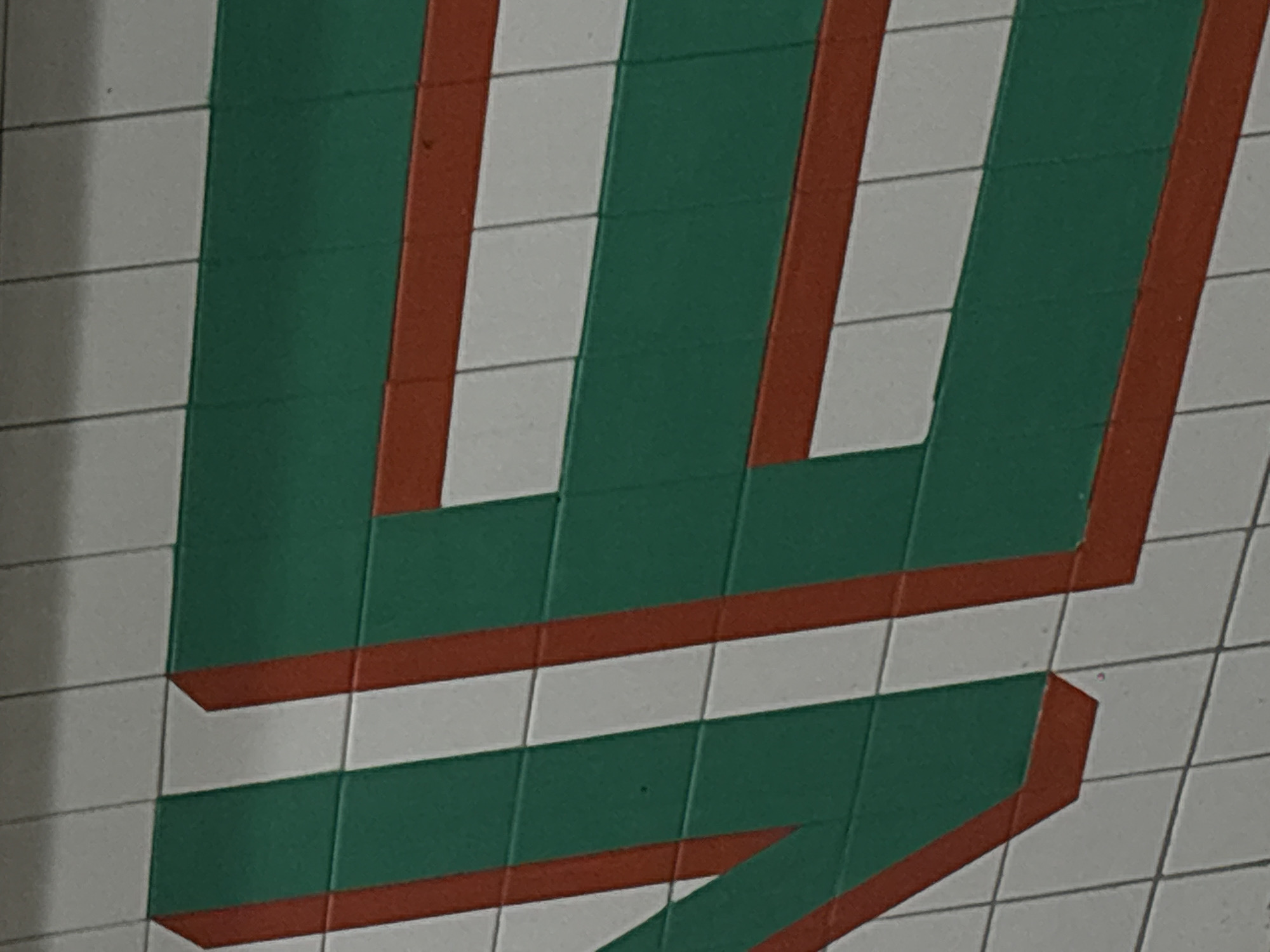 The image depicts a section of tiled wall featuring a green and orange letter "U" graphic, presumably part of the logo for the Universidad Iberoamericana, a Jesuit university in Mexico City, Mexico. The design consists of green stripes separated by orange outlines on a white tile background. The image exhibits an off-angle perspective, focusing primarily on the upper portion of the letter. The tiles appear clean and well-maintained, with visible grout lines.