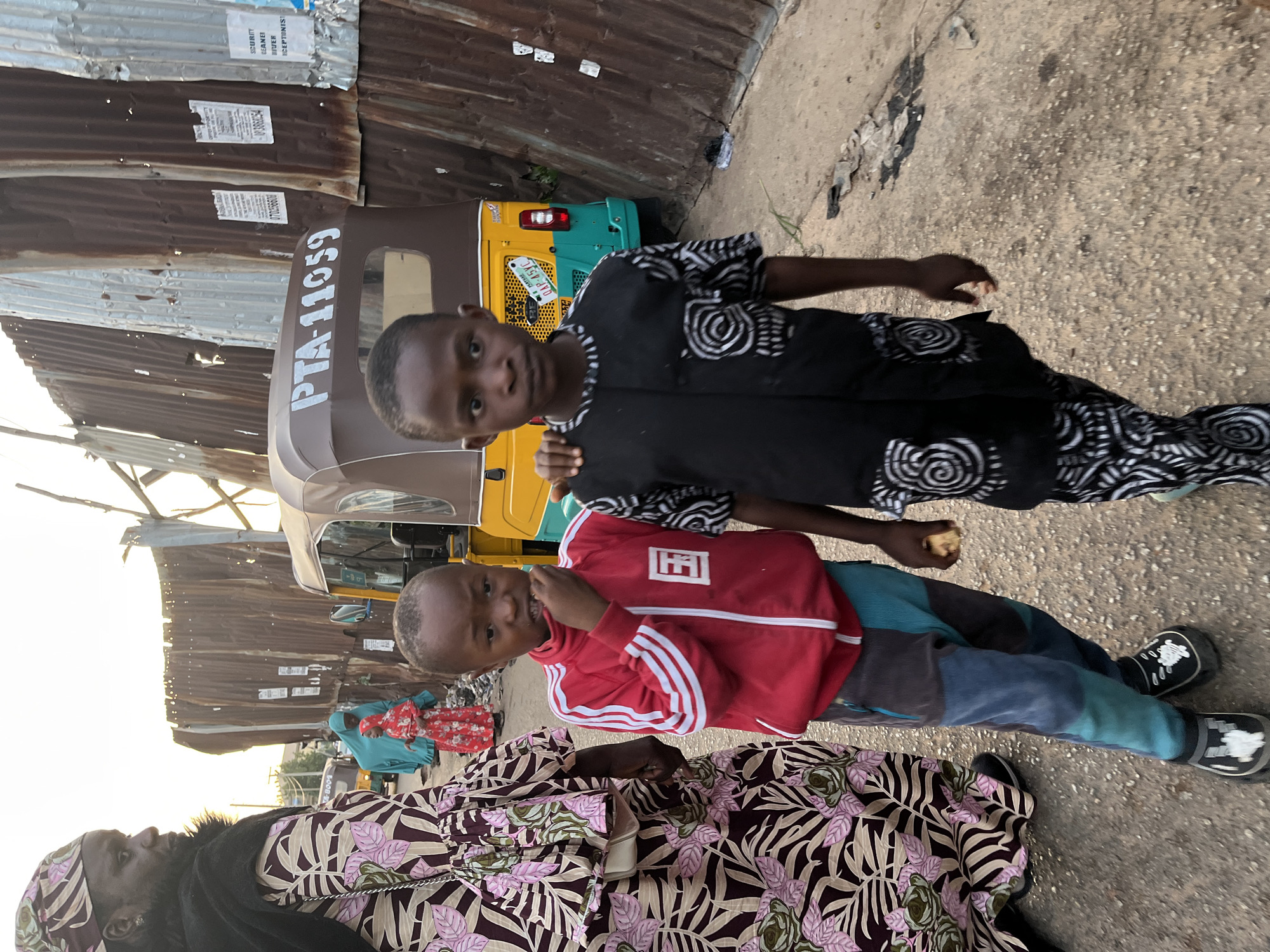Two young boys, one wearing a red track jacket with white stripes and colorful patchwork pants, the other in a black patterned tunic and pants, stand in a dirt street in Gura Top, Nigeria. The boy in red has his hand to his mouth, and the boy in black has his arm on the other boy's shoulder. Behind them, a yellow and teal auto-rickshaw is parked next to a corrugated metal wall adorned with posters. In the background, other individuals in traditional clothing can be seen. The overall scene depicts a street in a bustling urban environment.