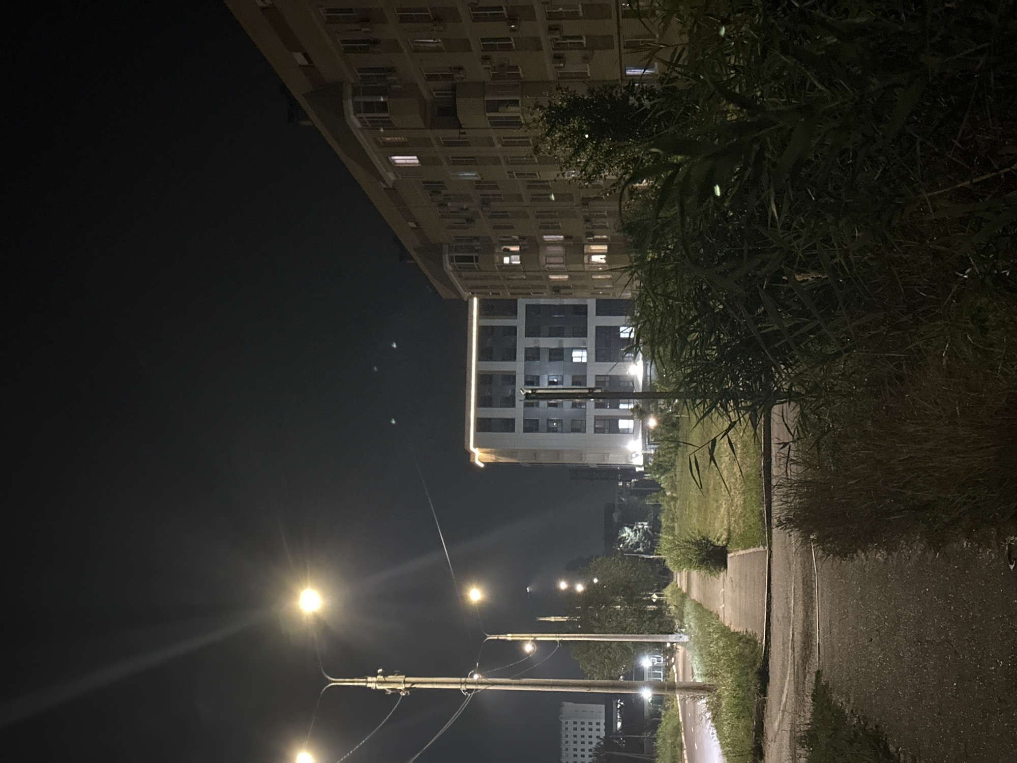 This is a nighttime, eye-level, medium shot of a street in the city of Nevinnomyssk, Russia. The image captures a sidewalk, a road, and several buildings illuminated by streetlights and interior lights. In the foreground, a paved road curves to the left, bordered by a grassy area and dense vegetation. A concrete sidewalk runs parallel to the road, leading into the distance.
Dominating the right side of the frame is a multi-story apartment building with a beige facade. Its windows are illuminated, indicating occupancy. To the left of this building, across the sidewalk, is another, more modern building with a white and grey facade, also lit from within. In the far background, another building with a lighter facade is visible.
Several streetlights are positioned along the sidewalk, casting a yellow glow. Power lines are visible, strung between utility poles. The sky is dark, characteristic of nighttime. The overall impression is that of a residential urban environment at night. There are no people visible in the image.