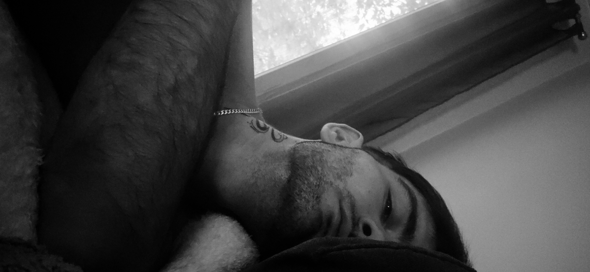 This black and white, close-up image features a person, likely male, lying down indoors. The frame is tilted, presenting the subject diagonally.
The primary subject is a person, whose side profile is visible, including a hairy arm, neck with a patterned tattoo, an ear, and the side of their face with facial hair (beard/stubble). Their eye is partially open, conveying a relaxed, sleepy, or contemplative expression. A chain-link bracelet is worn on their wrist or lower arm. The person appears to be resting on a dark, soft material, possibly a blanket or pillow.
In the background, a window is visible, letting in diffused light, suggesting a daytime indoor setting. A window frame or curtain rod runs along the top edge of the image. The overall atmosphere is quiet and intimate, enhanced by the monochromatic aesthetic. No visible text is present in the image.