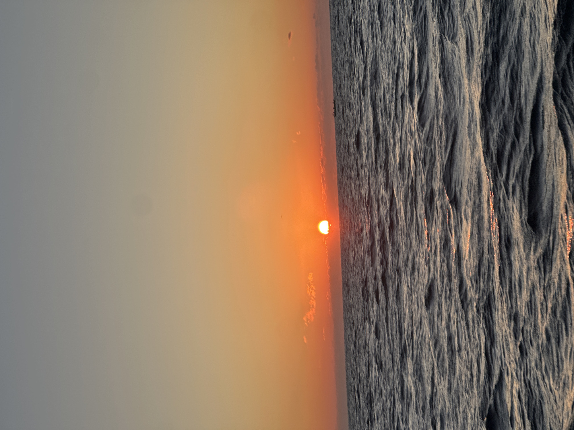 The image shows a sunset over the ocean. The sun is a bright orange ball on the horizon, casting a warm glow across the sky and water. The sky is a gradient of orange and yellow, with a few wispy clouds. The ocean is dark and choppy, with small waves reflecting the sunlight. In the distance, a small boat can be seen on the water. The location is unknown.
