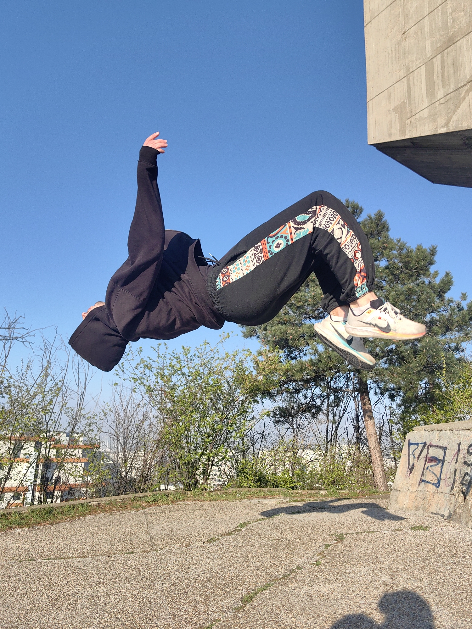 A person wearing a black hoodie and black sweatpants with a colorful patterned stripe down the side is in mid-air, performing a backflip. They are wearing off-white sneakers with a colorful translucent sole. The backdrop consists of a clear blue sky, trees, and a low building in the distance. A concrete structure is visible on the right side of the frame. The person's shadow is cast on the cracked concrete ground, near a graffiti-covered low wall. Based on the visual cues, the location is likely in Varna, Bulgaria.