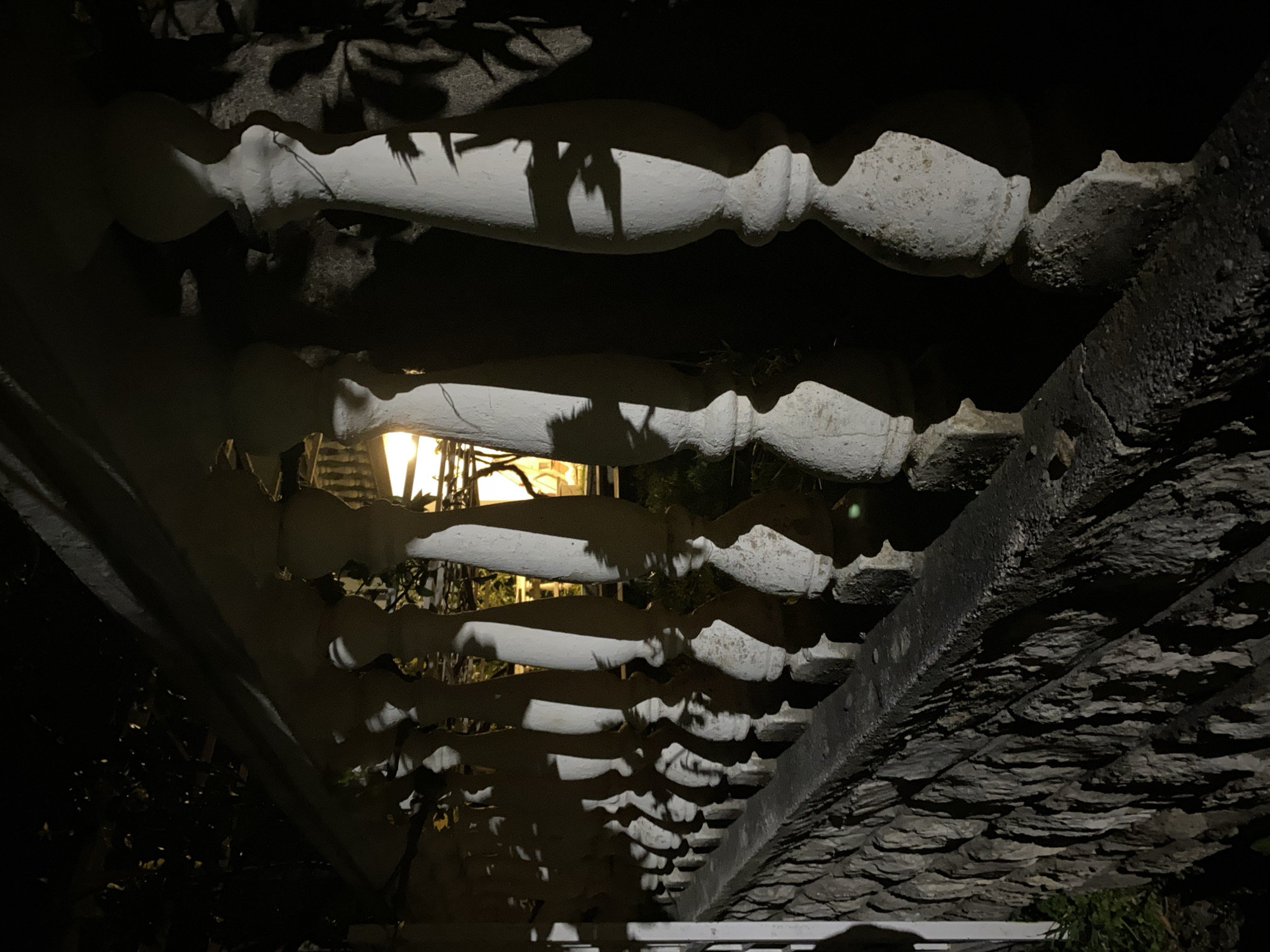 The image depicts a view under a stone balustrade in Budva, Montenegro, taken at night. The balusters are white and cast shadows onto the stone surface above. A light source is visible through the balustrade, illuminating the space beyond. The texture of the stone is rough and uneven. The overall lighting is dark with strong contrasts.