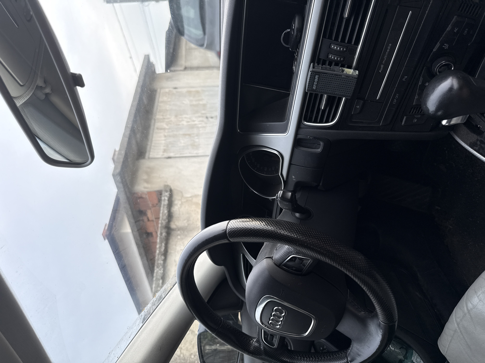 The image shows the interior of a car, likely an Audi, from the perspective of the driver. The steering wheel, with the Audi logo visible, is in the foreground. The dashboard features air vents, buttons labeled "ESP OFF," "P," and "AC OFF," and a multimedia system. An "areon" brand air freshener is clipped to an air vent. The rearview mirror is visible, reflecting an overcast sky and a building with a tiled roof, suggesting a cloudy day. The overall setting appears to be a parking area in Skutare, Bulgaria, given the location context provided. There is no clear indication of any activity or event taking place, other than potentially being parked. The mood is neutral, with no strong emotional cues conveyed. The image is taken from inside the car looking outwards and towards the dashboard.