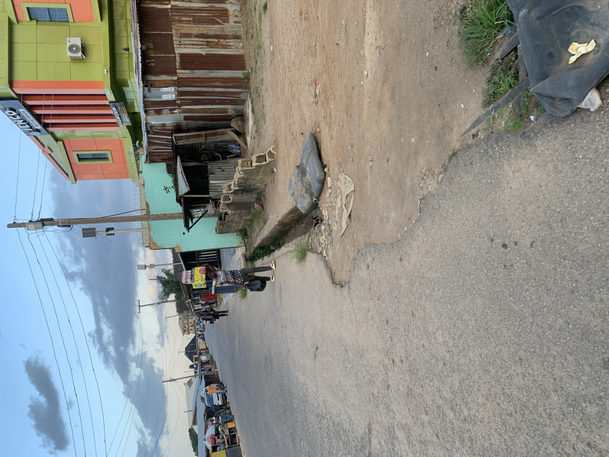 The image depicts a street scene in Jos, Nigeria. The road is paved and appears to be a busy thoroughfare, with a few vehicles, including a white van, visible in the distance. On the left side of the street, market stalls are set up under awnings, with people browsing and interacting. A woman is seen walking in the middle of the road, carrying a black bag and a tray on her head, loaded with goods. On the right side of the street, there are some makeshift structures made of corrugated iron, and a more modern building with a brightly colored facade featuring the text "JENCO MULTI-PLATE SERVICES". The sky is cloudy with hints of blue. A drainage ditch runs alongside the road on the right, with some debris and vegetation present. In the foreground, a dark, irregularly shaped object lies on the dusty ground.