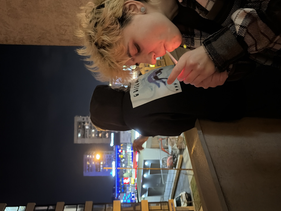 The image captures a nighttime urban scene from a balcony in Sofia, Bulgaria. In the immediate foreground, an individual with short, spiked, bleached blonde hair and a visible earlobe gauge is shown in profile, wearing a black, white, and grey plaid jacket. This individual is actively lighting a cigarette held in their mouth using a red-glowing lighter, with visible exhalation of smoke. Behind this individual, a second person is partially visible, wearing a dark hooded sweatshirt featuring a white graphic print on the back. This second individual stands facing away from the camera, looking out over the illuminated cityscape.
The environment below the balcony reveals a street with various light sources, including blue and red commercial or traffic lights. A white van is parked on the street level, adjacent to what appears to be a construction zone or an undeveloped area containing an orange excavator. In the mid-ground and distant background, multiple multi-story buildings are illuminated, silhouetted against a dark night sky. The balcony itself features a concrete or painted railing in the lower foreground.