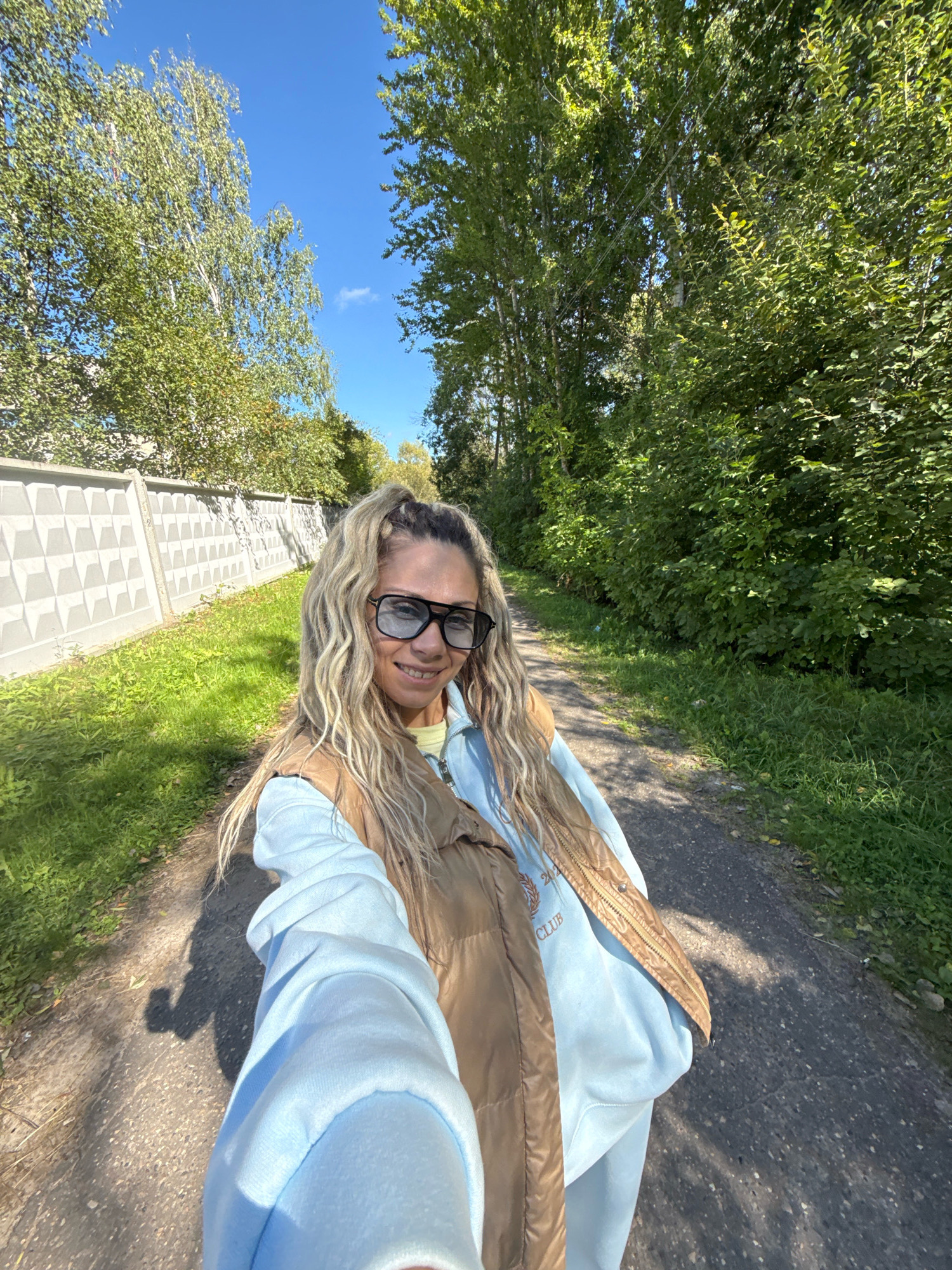 An outdoor, eye-level, medium selfie shot shows a woman with blonde, curly hair pulled into a ponytail, wearing dark sunglasses, a light blue hoodie, and a brown puffer vest. She is smiling at the camera, holding the camera to take the selfie. The woman is walking on a paved path bordered by grass and trees. To her left is a grey, textured concrete fence. The scene appears to be in Rusanovo, Russia, on a clear, sunny day.