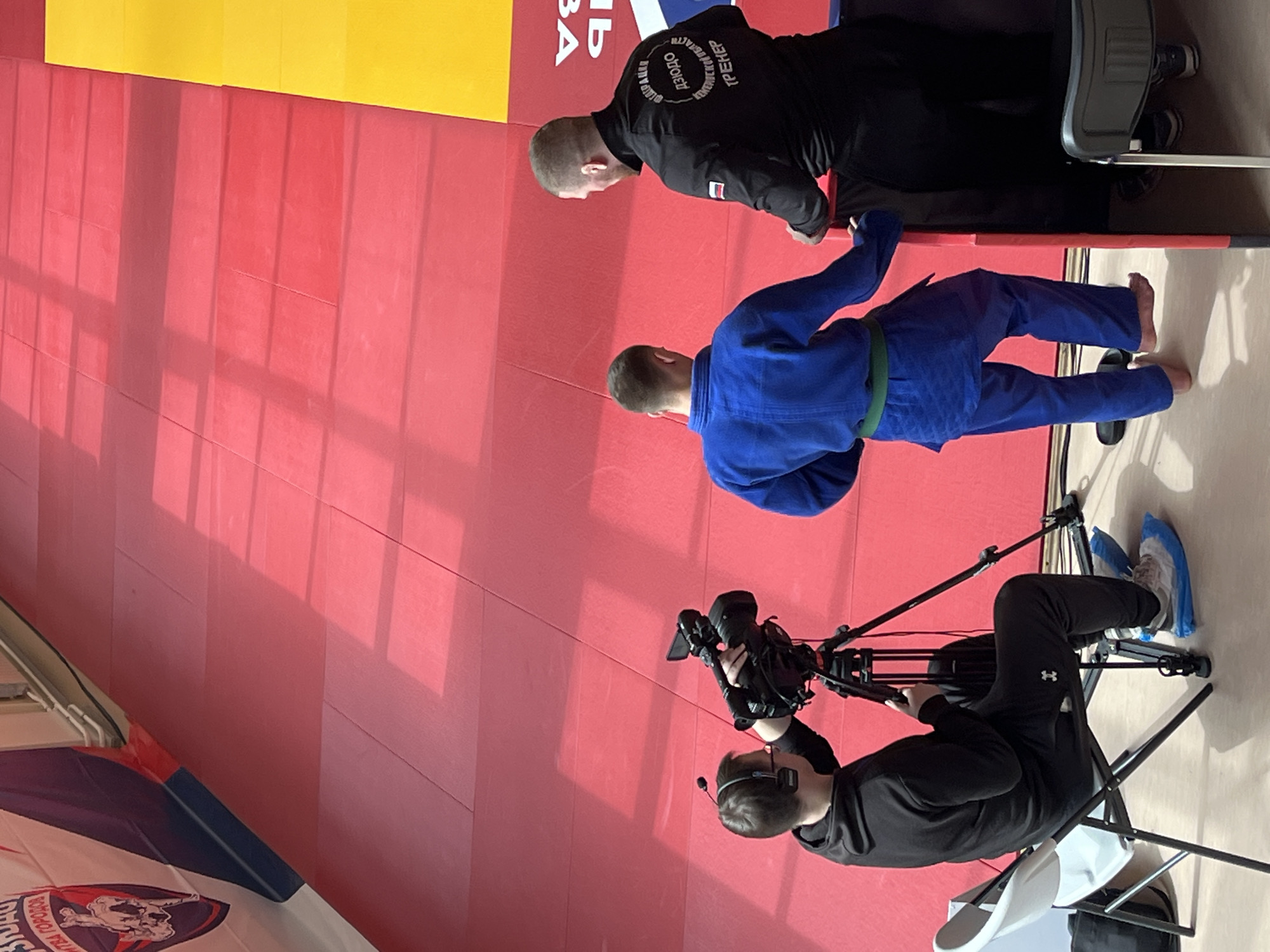 The image shows a martial arts event, likely a competition or demonstration, in Krasnoyarsk, Russia.
Two male martial artists stand on a red and yellow padded floor. One wears a black uniform, and the other wears a blue uniform with a green belt. The black-uniformed individual appears to be either a trainer or a more experienced competitor. A camera operator sits on a chair, filming the scene.
The background features a red padded wall. In the lower-left corner, a flag with Cyrillic text is visible. The cameraman has a headset on and is operating a professional video camera mounted on a tripod. He is wearing shoe covers.