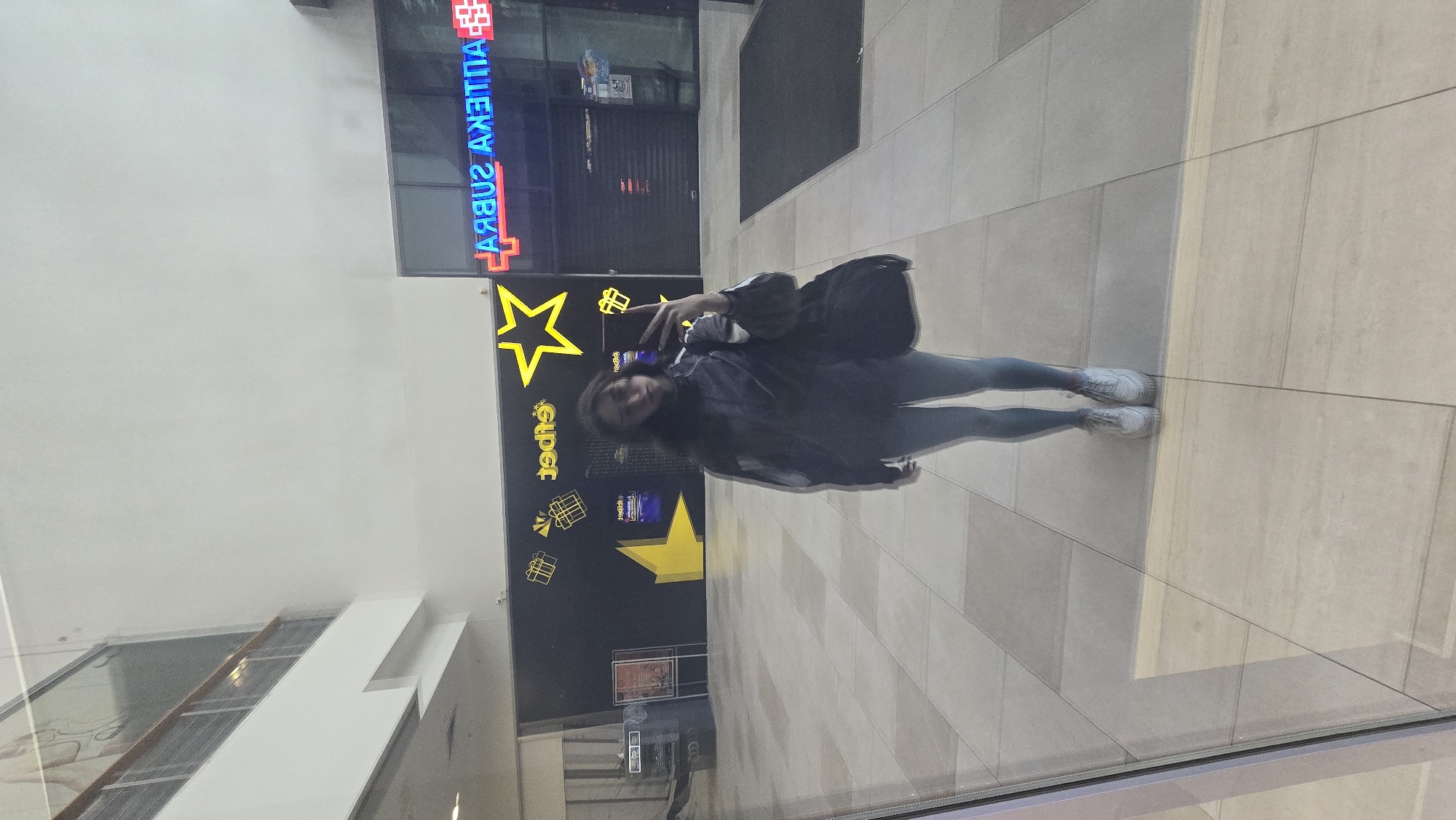 The image, rotated, depicts a young woman standing indoors, likely within a shopping mall or commercial complex in Stara Zagora, Bulgaria. She is positioned on a light-colored tiled floor, against a backdrop of a plain white wall and a dark wall adorned with bright yellow star and gift box graphics.
The woman is wearing a dark jacket or hoodie, grey leggings, and white sneakers. A dark shoulder bag hangs from her left shoulder. She is holding a smartphone in her right hand, possibly taking a selfie or viewing the screen, and looks towards the camera with a neutral to slightly smiling expression.
Behind her, on the dark wall, prominent yellow neon-like graphics include stars and gift boxes, along with the word "Super" (with the 'i' stylized as a star). In the upper background, through a glass facade or window, neon signage is visible. The text appears in reverse due to reflection, clearly spelling "APTЕКА SУБРА" (APTEKA SUBRA) in red and blue neon, indicating a pharmacy. A glass barrier or window is present in the immediate foreground, causing reflections on the floor. The overall setting is well-lit, typical of an indoor commercial environment.