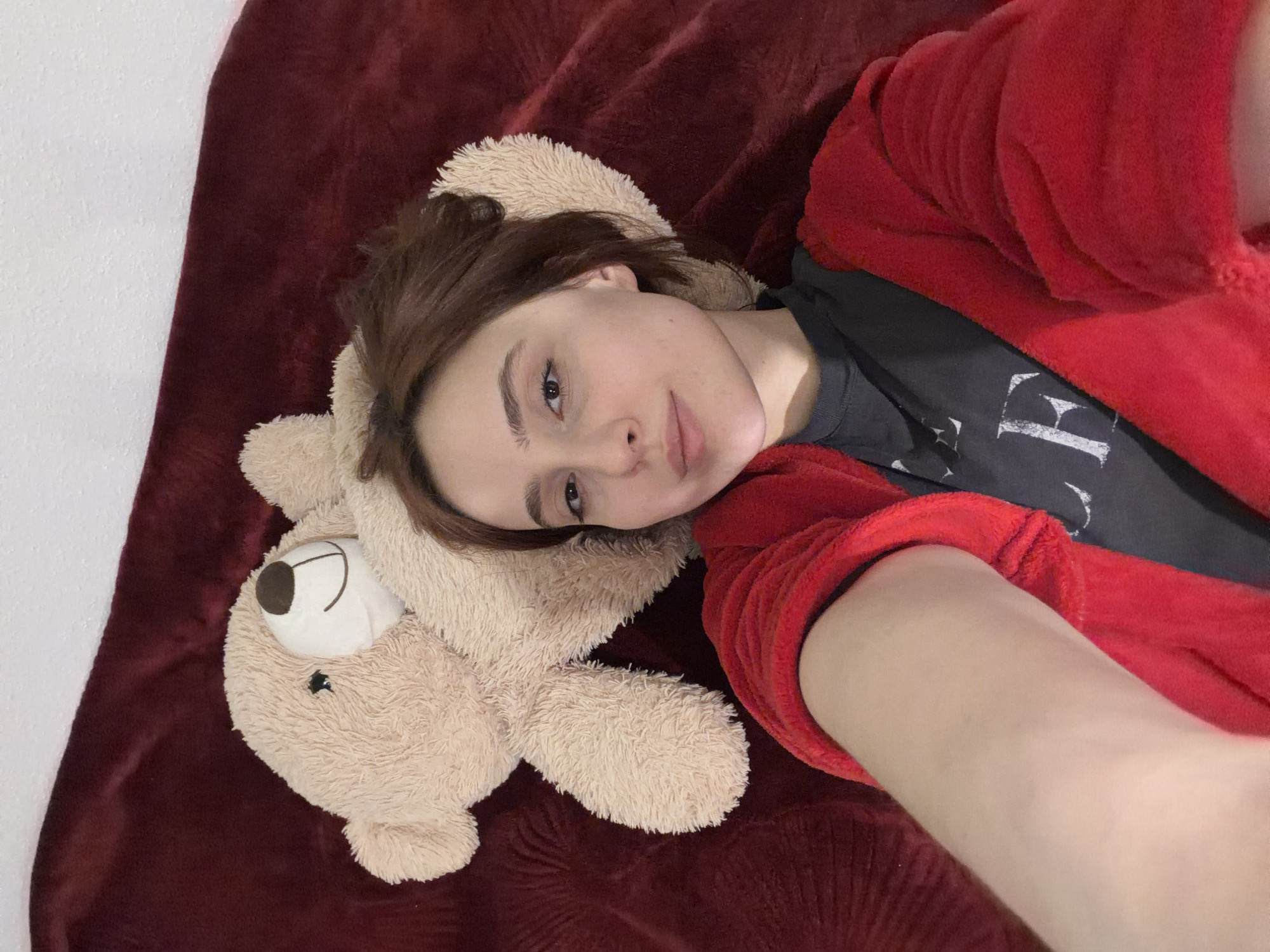 The image displays a young female subject and a teddy bear in an indoor setting. The female, with light skin, brown hair, and brown eyes, is lying down, positioned centrally, looking directly into the camera with a neutral expression. She is wearing a dark grey t-shirt, partially visible under a red, textured robe or blanket that covers her upper body. Her right arm extends towards the camera, indicating she is holding the device to capture a selfie. A light brown, fluffy teddy bear with dark eyes and nose is situated to the left of her head, resting on the same surface. The immediate environment consists of a dark red, textured blanket or comforter, likely covering a bed. A segment of a plain white wall is visible in the upper left background. The location in the city of Strasbourg, France, cannot be determined from the visual information present in the image.