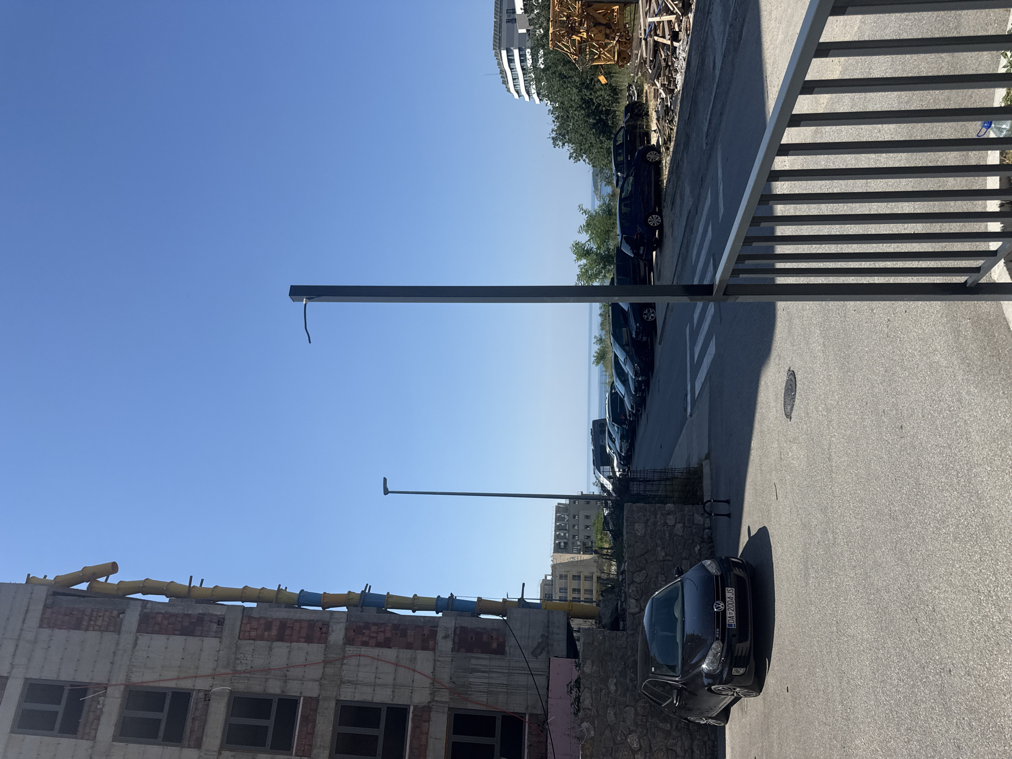 This media file captures a scene in Boreti, Montenegro, on a clear, sunny day. The foreground features a construction site with a partially built brick and concrete structure, equipped with yellow and blue tubular scaffolding. To the right, a gray metal railing overlooks a paved street where several dark-colored cars are parked. One car, a black Volkswagen Golf GTI, is prominently visible with its license plate reading "DA 2004 JS". Further down the street, more cars are lined up, and beyond them, lush green trees and distant buildings can be seen. The sky is a vibrant blue, suggesting it is daytime, likely late morning or afternoon given the shadows cast. The overall impression is one of ongoing development and a typical Mediterranean coastal town.