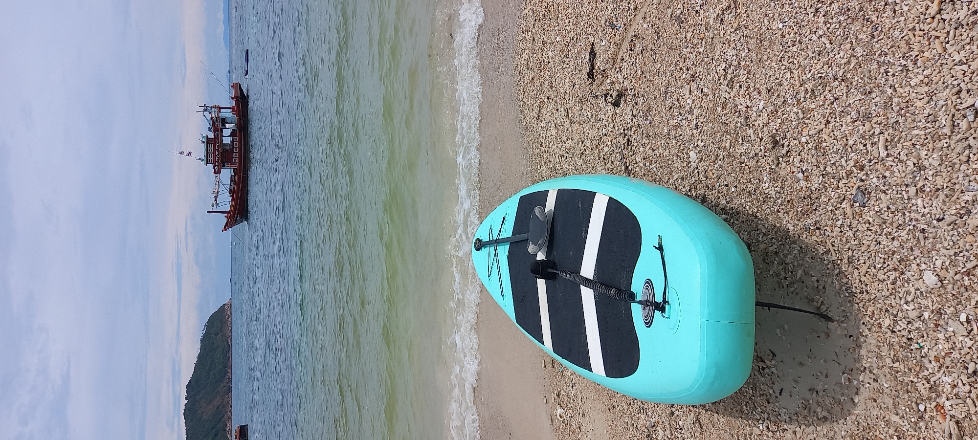 The scene is a beach in Baan Chaloklum, Thailand. A light blue paddleboard lies on the sandy beach close to the water's edge. A paddle is resting on top of the board. A boat is in the water, and land is visible in the background. The sky is overcast.