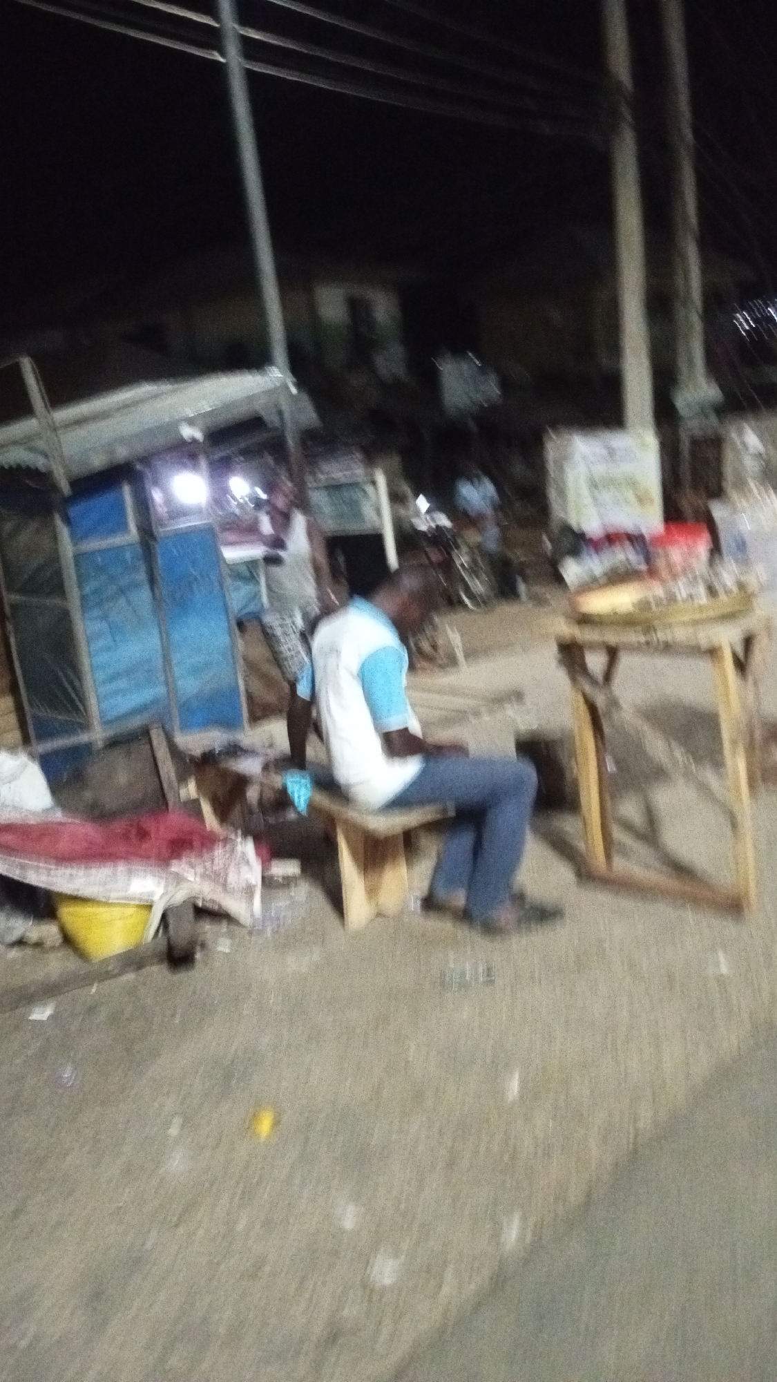 The media file presents a blurry, low-light image of a street scene at night in Adankolo, Nigeria. The setting appears to be an informal commercial area, possibly near market stalls or roadside shops, illuminated by artificial light sources.
Two men are the primary subjects. In the foreground, a man with dark skin is seated on a low, wooden bench. He is wearing a white t-shirt with light blue sleeves and blue jeans, with dark slip-on shoes on his feet. He appears to be looking downwards or slightly to his right. Behind him and to his left, another man with dark skin is standing, wearing a light-colored top and patterned shorts. He is partially obscured by the foreground elements.
To the left, a makeshift stall or shack with blue and brown panels is visible, with a bright light source illuminating its immediate area. To the right of the seated man, there is a simple wooden table holding various indistinct items, possibly goods for sale. In the left foreground, a red fabric and a yellow bucket are on the ground. Faintly in the background, a bicycle can be seen. Above, power lines stretch across the dark sky, supported by utility poles. The ground beneath appears to be unpaved, possibly dirt or rough concrete, with some scattered debris.
The scene depicts a quiet moment, likely an evening rest or wait. The overall atmosphere suggests an everyday, lived-in environment. Due to the blurriness and low light, no discernible text is visible on any signs or objects.