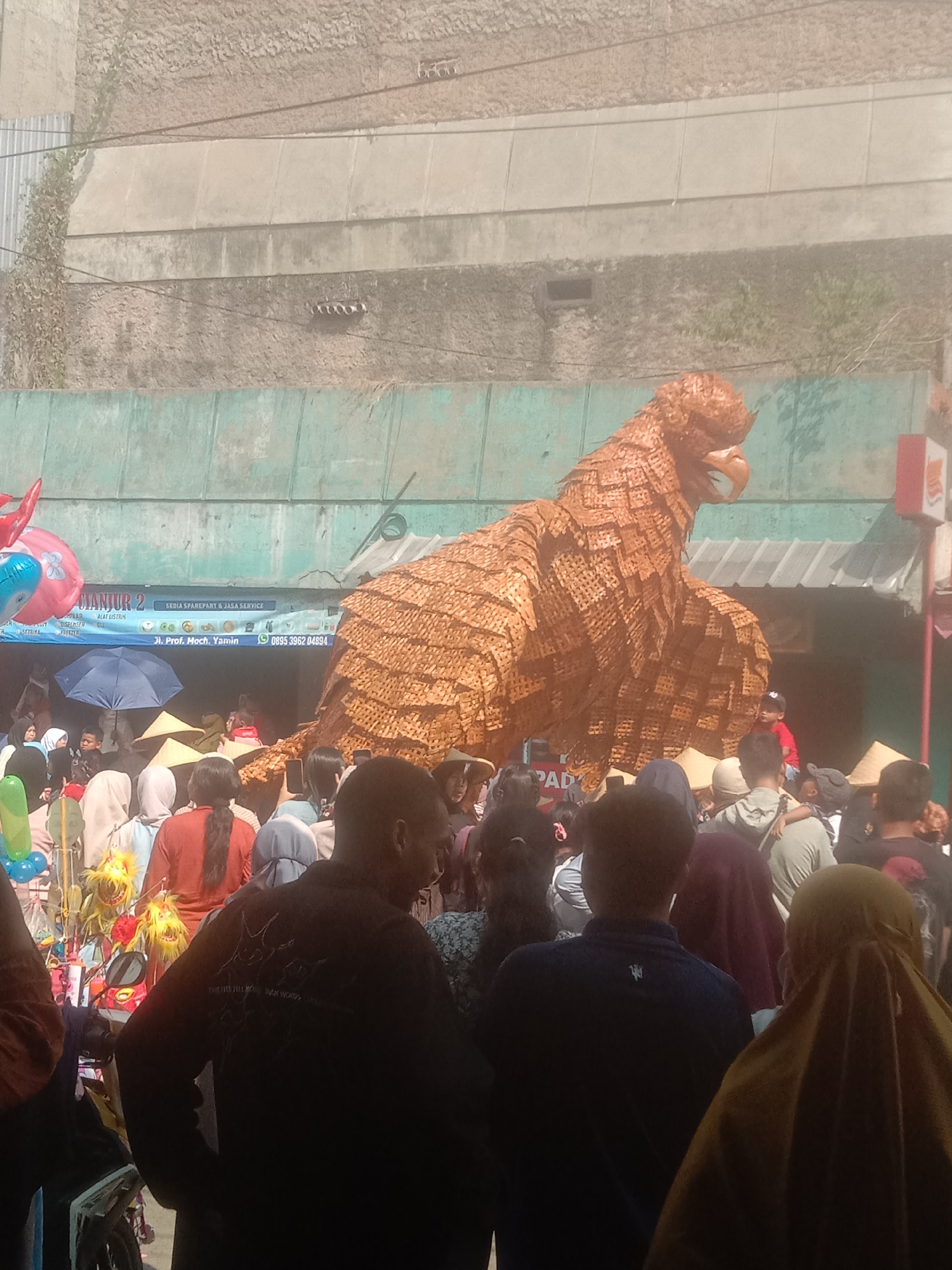 This is a photograph taken outdoors, likely in the city of Cianjur, Indonesia, given the presence of a sign that mentions "CIANJUR 2". The scene depicts a crowded street or open area with a large, woven effigy of a bird of prey, possibly an eagle, as the central subject. The effigy is golden brown and appears to be constructed from woven plant material. It is positioned in front of a building with a green facade and a textured, beige upper wall.
In the foreground and midground, a diverse crowd of people are visible. Many are wearing casual clothing, and several individuals are wearing headscarves. Some people are looking towards the effigy, while others are engaged in conversation or interacting with their surroundings. There are also stalls or vendors present, with balloons and other items on display. One stall has a sign that reads "DIANJUR 2 SEDIA SPAREPART & JASA SERVICE" with additional text indicating an address and phone number. A blue umbrella is open among the crowd, providing shade. The overall atmosphere suggests a public gathering, possibly a festival or event. The lighting indicates it is daytime, with bright sunlight casting shadows.