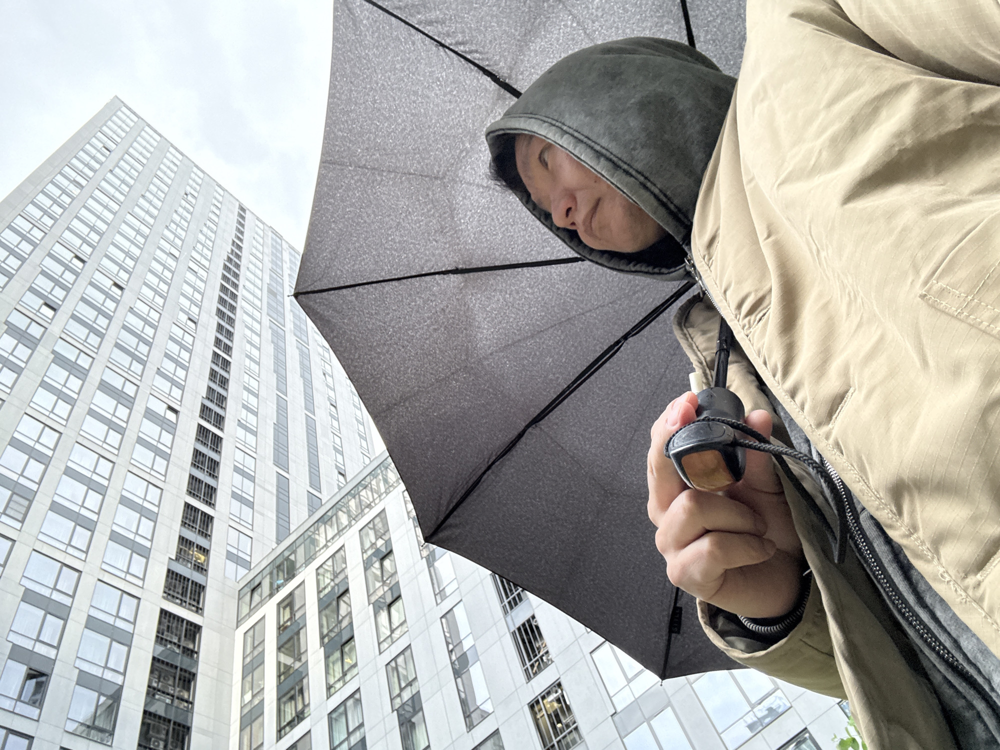 A low-angle shot depicts a person holding a closed umbrella, likely for protection from rain. The person is wearing a hooded jacket, the hood pulled up to cover their head. The umbrella is gray with dark ribs. In the background, two tall buildings are visible, their facades covered in windows. The sky is overcast. This scene appears to be set in a metropolitan environment, potentially in Moscow, Russia.
