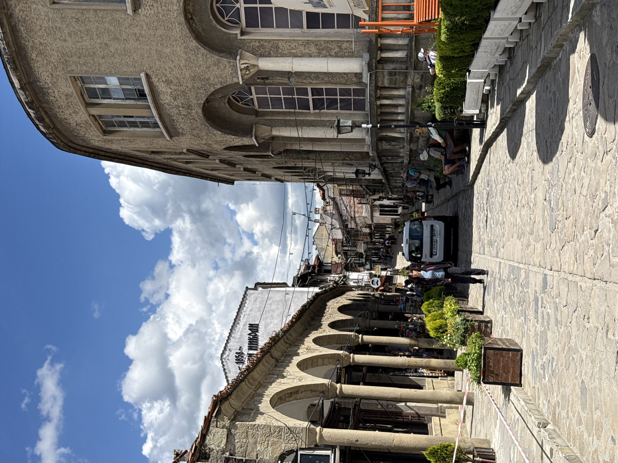 The image depicts a narrow street in Veliko Tarnovo, Bulgaria, characterized by historical architecture. A white van is positioned centrally in the street, heading away from the viewer. People are seen walking along both sides of the street and seated along the right. The buildings on either side of the street have stone facades with arched openings supported by columns, creating covered walkways. One building has the date 1858 inscribed on its facade. The street is paved with cobblestones. The sky above is blue with scattered clouds.