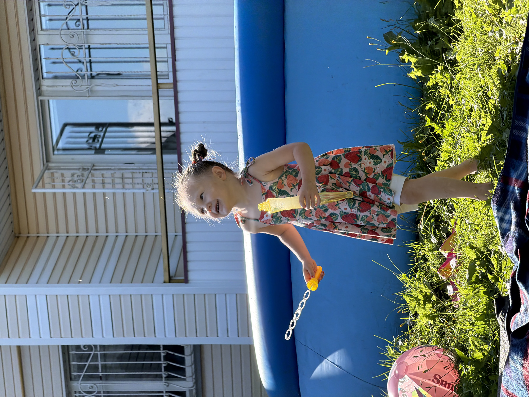 A young girl with light brown hair tied in two pigtails, wearing a pink and white floral dress and white shorts, is standing in a grassy yard next to a blue inflatable pool. She is holding a yellow bubble wand with multiple loops and a yellow bottle of bubble solution. The girl is looking up and smiling, appearing to be playing with bubbles. A pink inflatable ball is on the grass to her left, and a blue and red striped blanket is in the foreground. The background shows a two-story building with beige siding and white trim, featuring windows with decorative metal grates. The scene appears to be set in the city of Chaikovsky, Russia, on a sunny day.