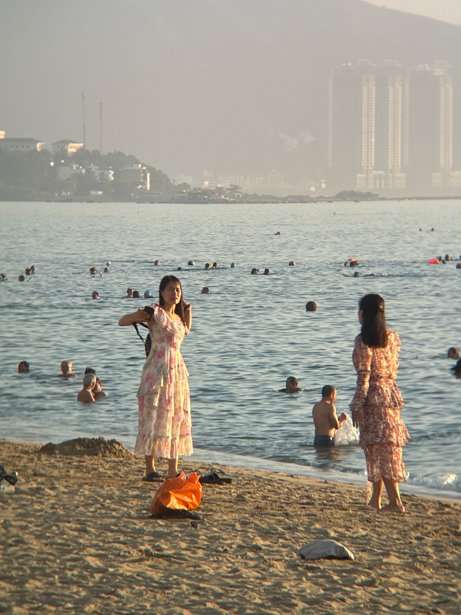The image depicts a beach scene in Nha Trang, Vietnam, with a hazy backdrop of distant buildings and mountains. In the foreground, two women stand on the sandy shore. The woman on the left wears a floral print dress and holds a phone. The woman on the right is also in a floral dress and faces away from the camera. Several people are visible in the shallow water, some swimming and others standing. An orange plastic bag lies on the sand near the woman on the left. The sea is calm and reflects the soft light. The overall atmosphere is one of a casual day at the beach.