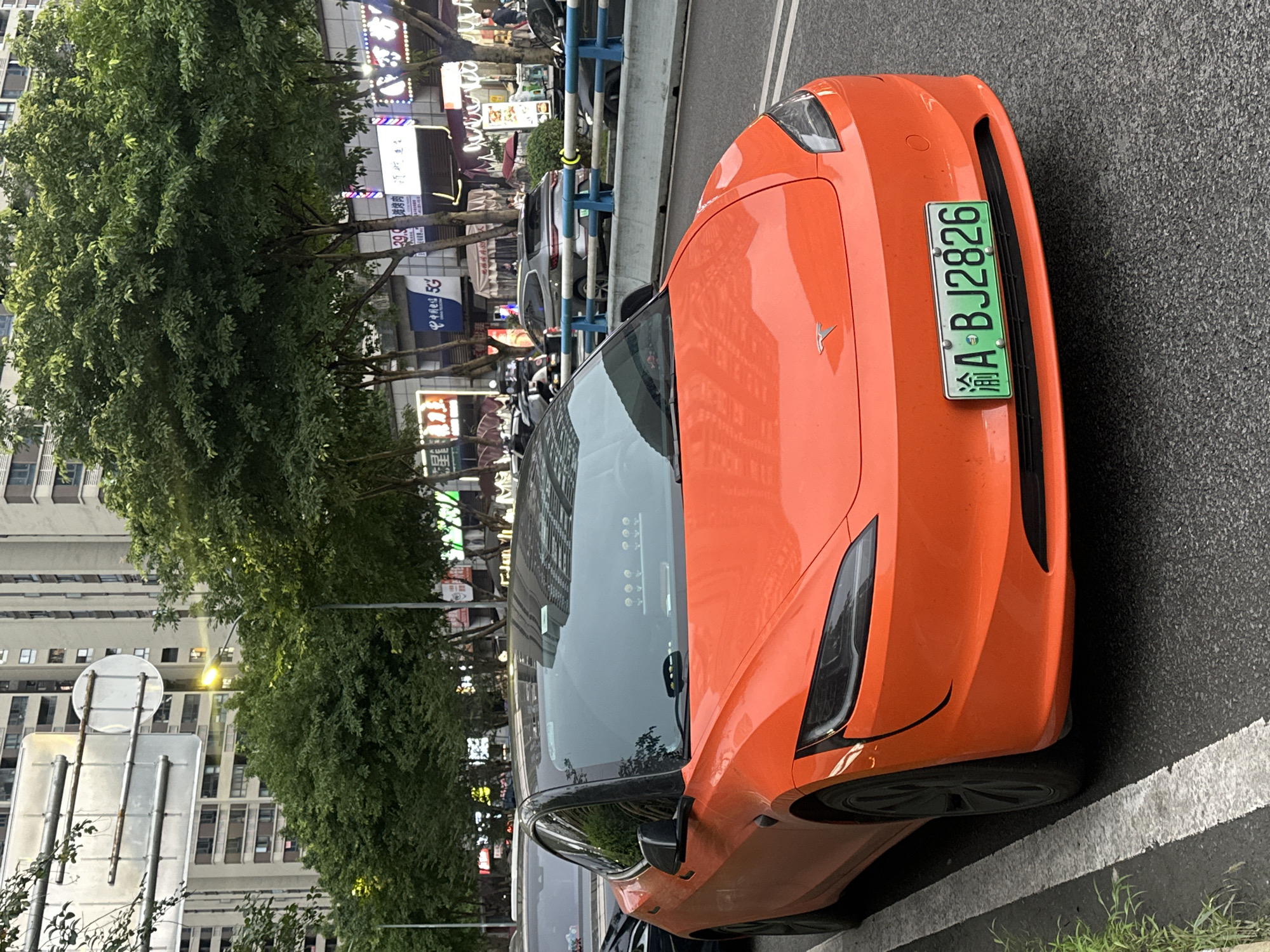 An orange Tesla is parked on the side of a street, with its license plate clearly visible. The license plate is green and displays the characters "渝A·BJ2826". The car is parked on asphalt, with a white line marking the edge of the parking area. In the background, there are trees and buildings, suggesting an urban setting. The buildings are multi-story and appear to be commercial or residential. Neon signs are visible on some of the buildings, indicating that it might be evening or nighttime, or that the area is well-lit. The overall scene is vibrant and dynamic, with the bright orange of the car contrasting with the green of the license plate and the foliage. The image appears to have been taken in Chongqing High-tech Zone, China, as indicated by the "渝" character on the license plate, which represents Chongqing. The Tesla's distinctive logo is visible on the hood. The lighting suggests it could be dusk or a moderately lit day, as shadows are not harsh. The car is the primary subject, but the surrounding urban environment provides context.
