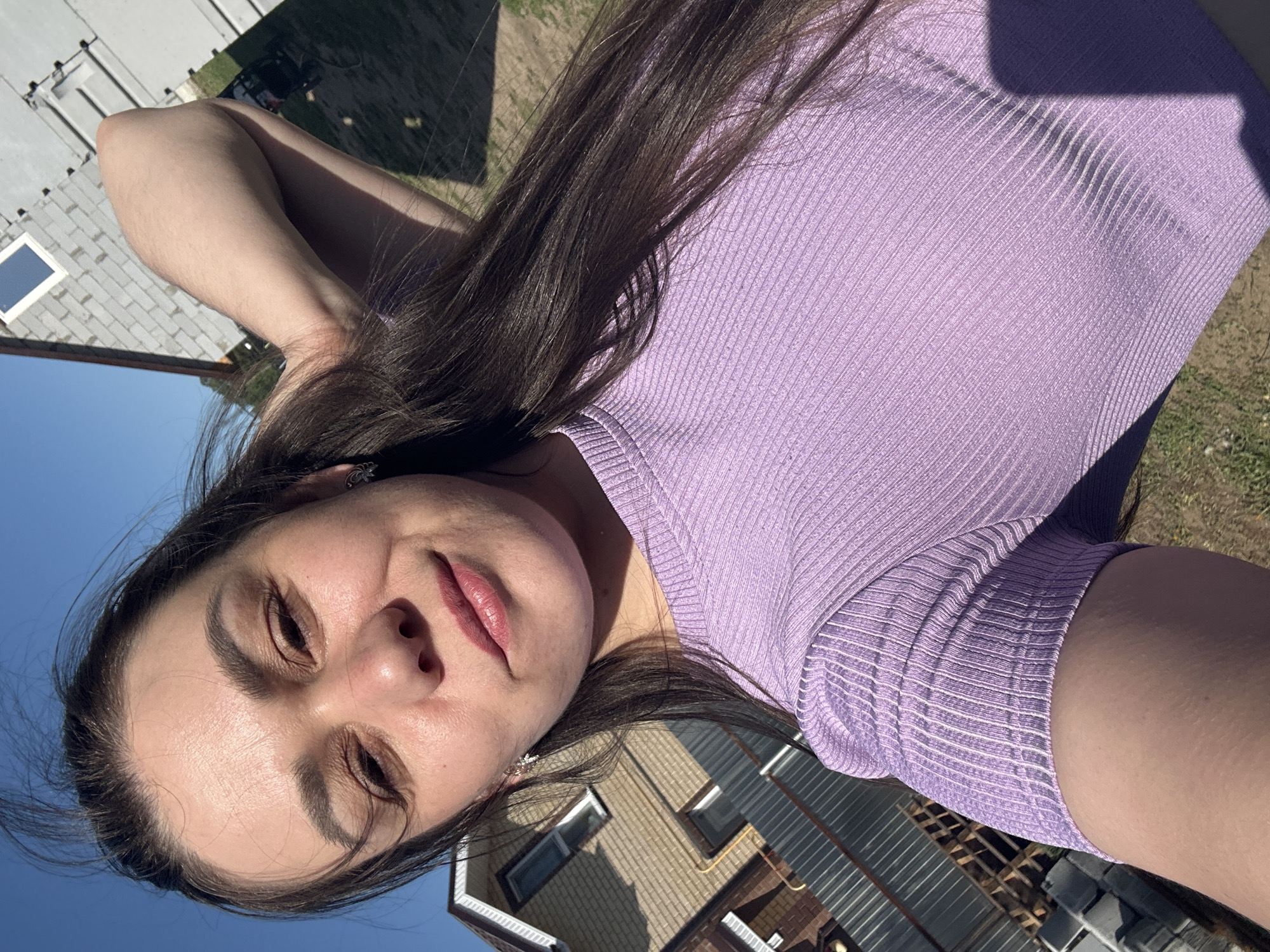 A woman with long dark hair is taking a selfie outdoors. She is wearing a ribbed purple top. The background shows a house with a beige exterior and white windows. The sky is blue. The image appears to be taken in Chaikovsky, Russia, based on the prompt.