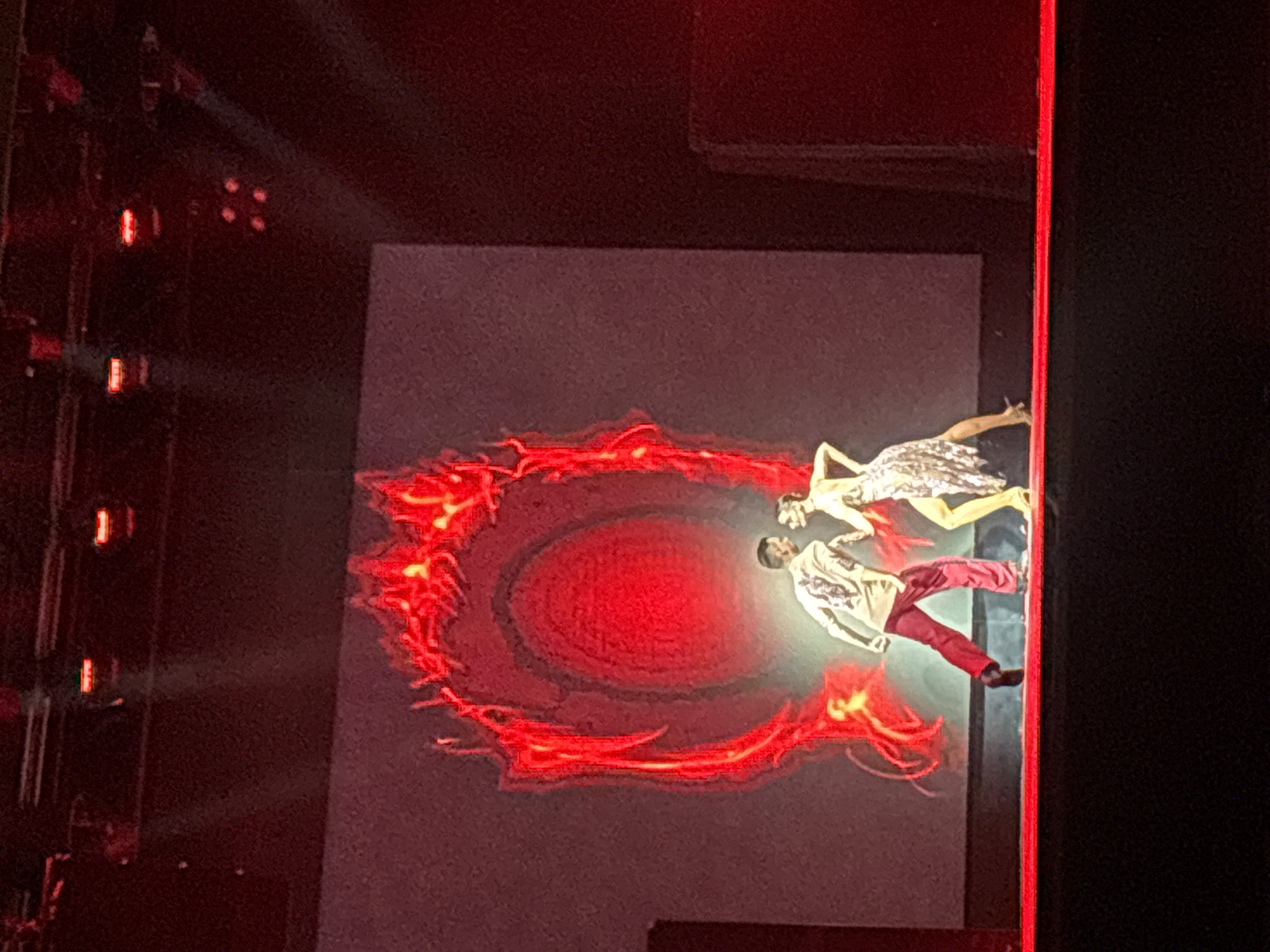 A man and a woman are dancing on a stage in front of a large screen displaying a fiery, red, oval-shaped graphic. The man is wearing a light-colored shirt with a dark pattern and red pants. The woman is wearing a shimmering, silver, short dress. They are facing each other and appear to be in motion. The stage is lit with red spotlights, creating a dramatic effect. The background is dark, with some architectural elements visible on the sides. The overall scene suggests a performance or show. The location is Unknown, Unknown.