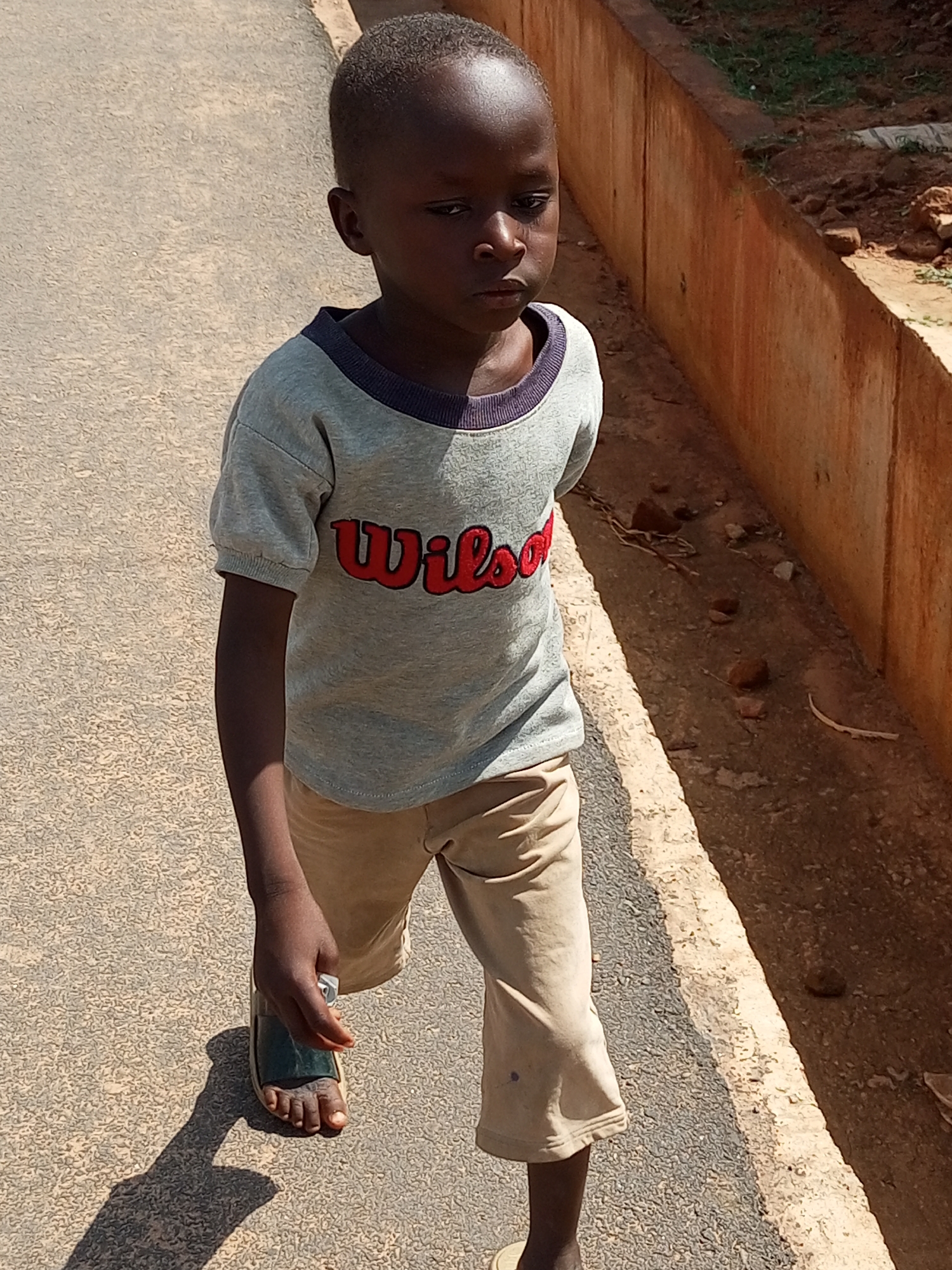 A young boy with dark skin and short hair walks along a paved path. He is wearing a light grey t-shirt with a navy blue collar and the red text "Wilson" in a cursive font across the chest. He also wears light tan pants that end just above his ankles. On his left foot, he wears a dark green and tan sandal. His right foot is bare. The boy is casting a long shadow on the path, suggesting it is either early morning or late afternoon. To his right, a red, concrete wall runs parallel to the path, with a patch of dry grass and soil visible beyond it. The scene appears to be outdoors in a sunny climate. The boy's expression is solemn, with his gaze directed downward. The ground is a textured, light-colored asphalt.