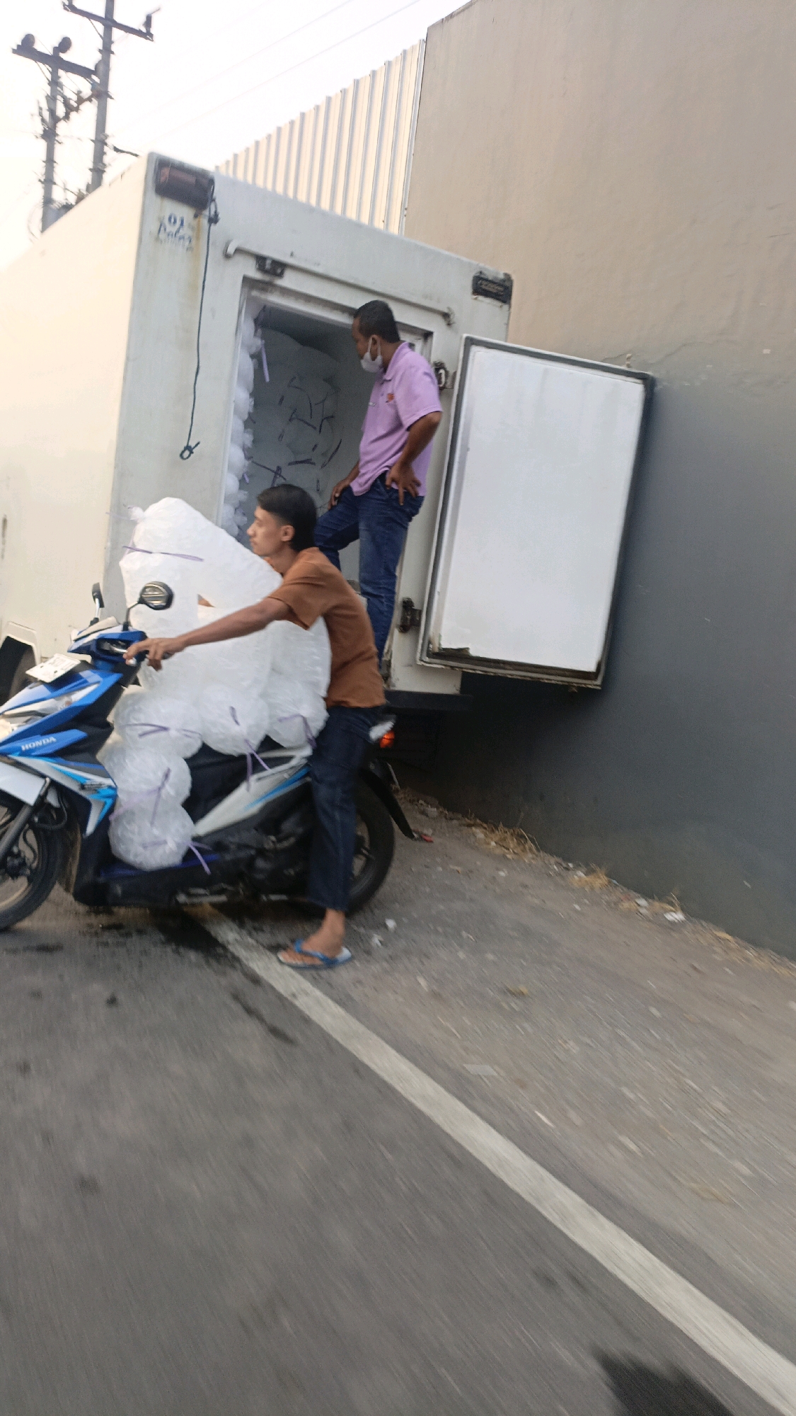 In the city of Salatiga, Indonesia, a man is seen on a motorcycle loaded with large, clear plastic bags filled with ice. He is wearing a brown t-shirt, dark pants, and blue flip-flops. Beside him, a white refrigerated truck is parked, its rear door open. Another man, wearing a pink shirt and blue jeans, is standing inside the truck, appearing to be working. The truck is parked on a street with a white painted line. The scene is outdoors with a cloudy sky and a building with a corrugated metal roof in the background.